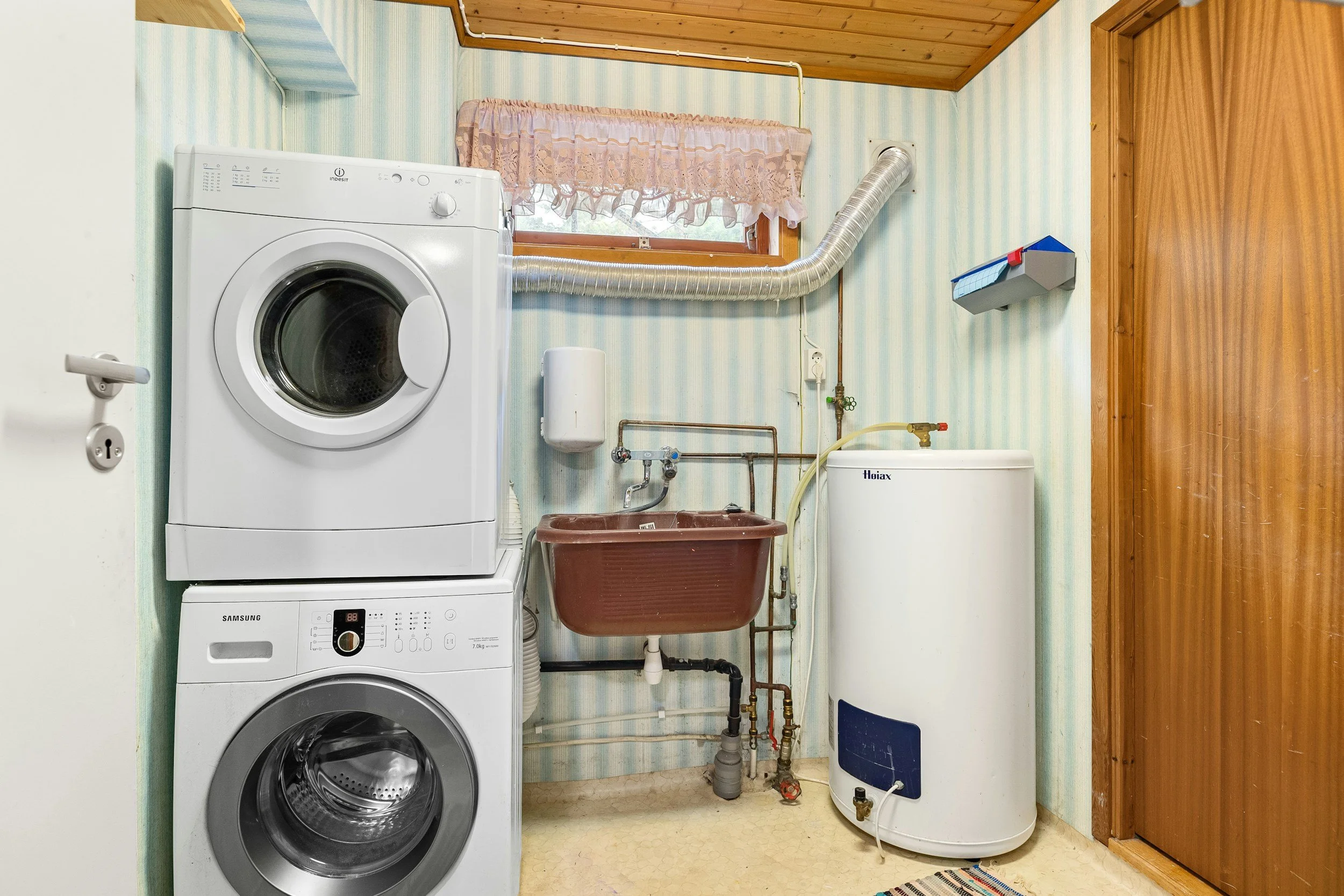 Laundry room with stacked washer and dryer, utility sink, water heater, small window with pink curtain, and various pipes and plumbing fixtures.