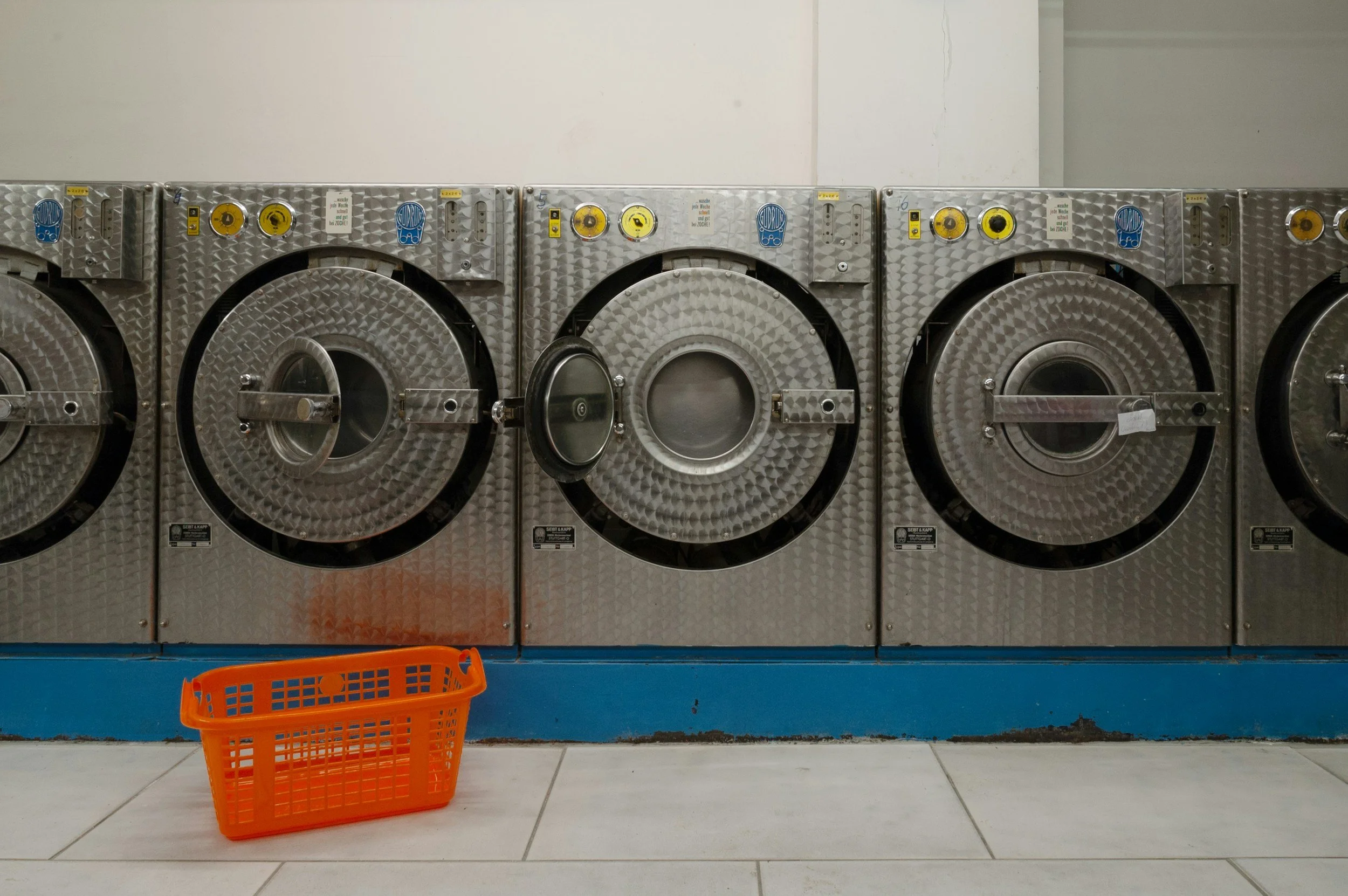 Laundry room with four front-loading industrial washing machines, an orange plastic laundry basket on the floor in front of them, white tiled floor, and a plain white wall.