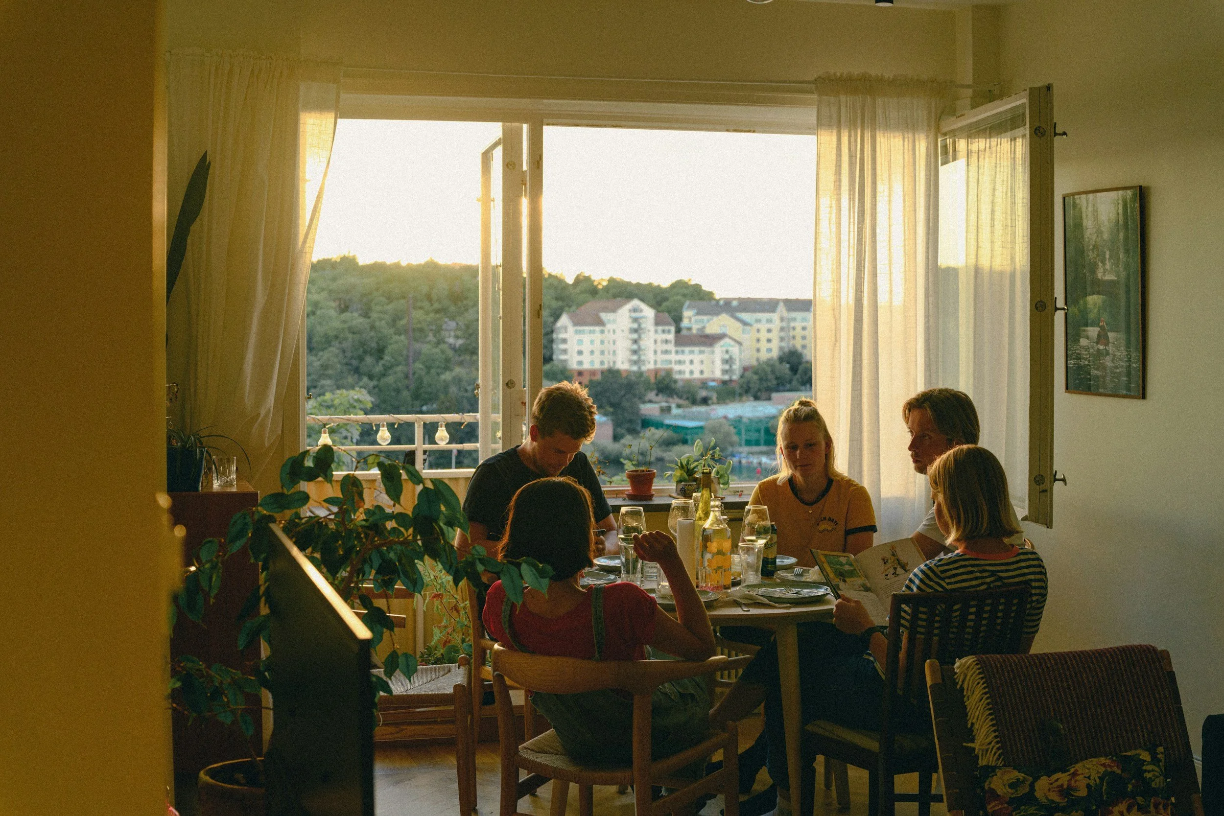 A family gathered around a dining table inside a home, with sunlight coming through open windows revealing a view of buildings and greenery outside.