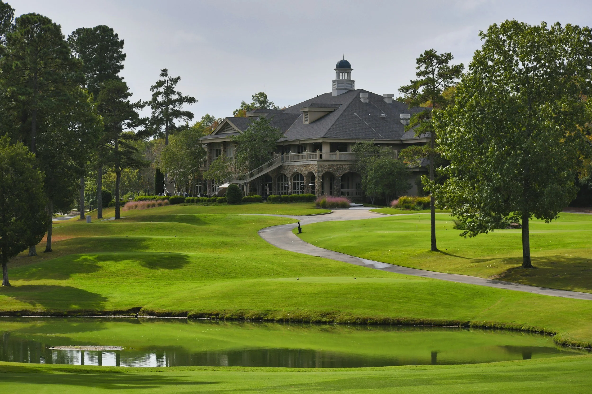 A large house with a dark roof is situated on a golf course with green grass, trees, and a small pond in the foreground.