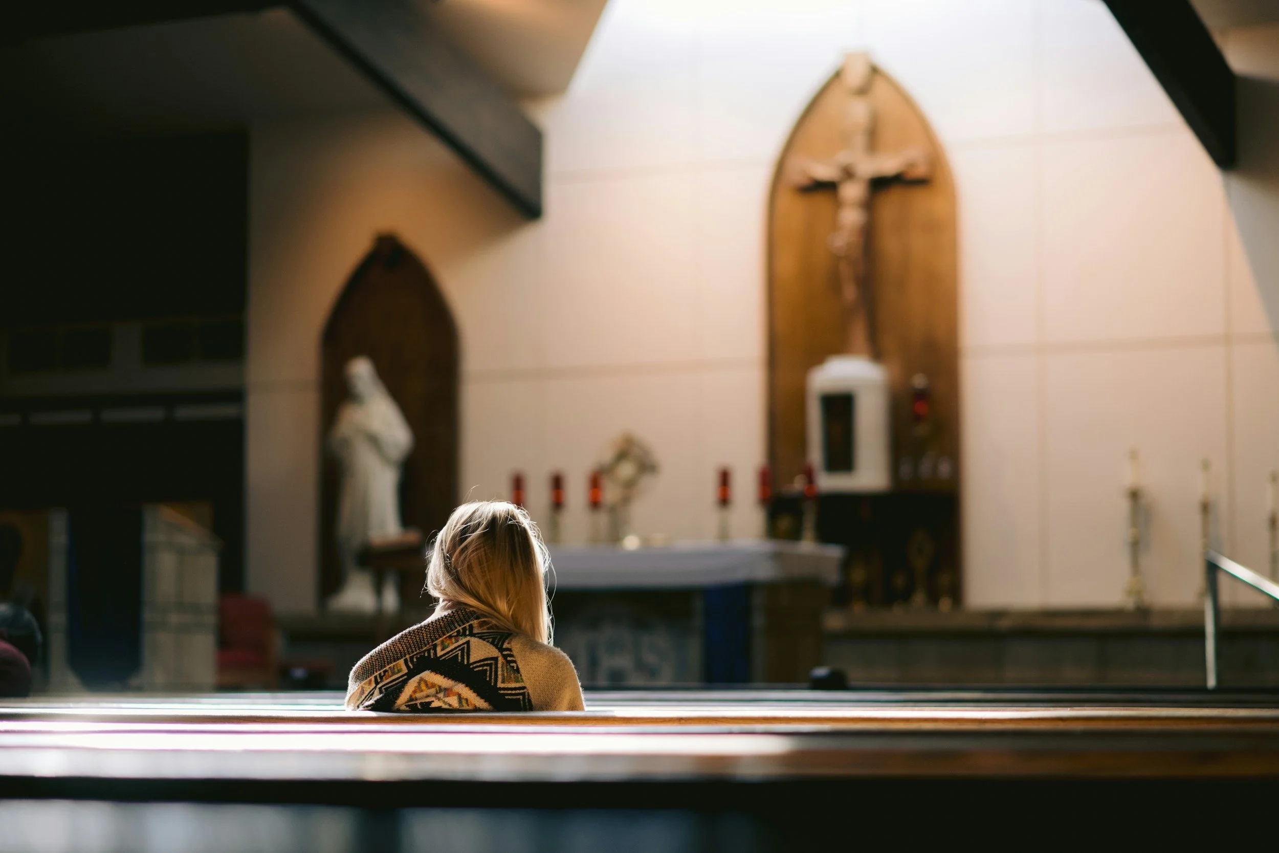 A woman sitting alone in a church pew, facing the altar which includes religious statues and a crucifix.