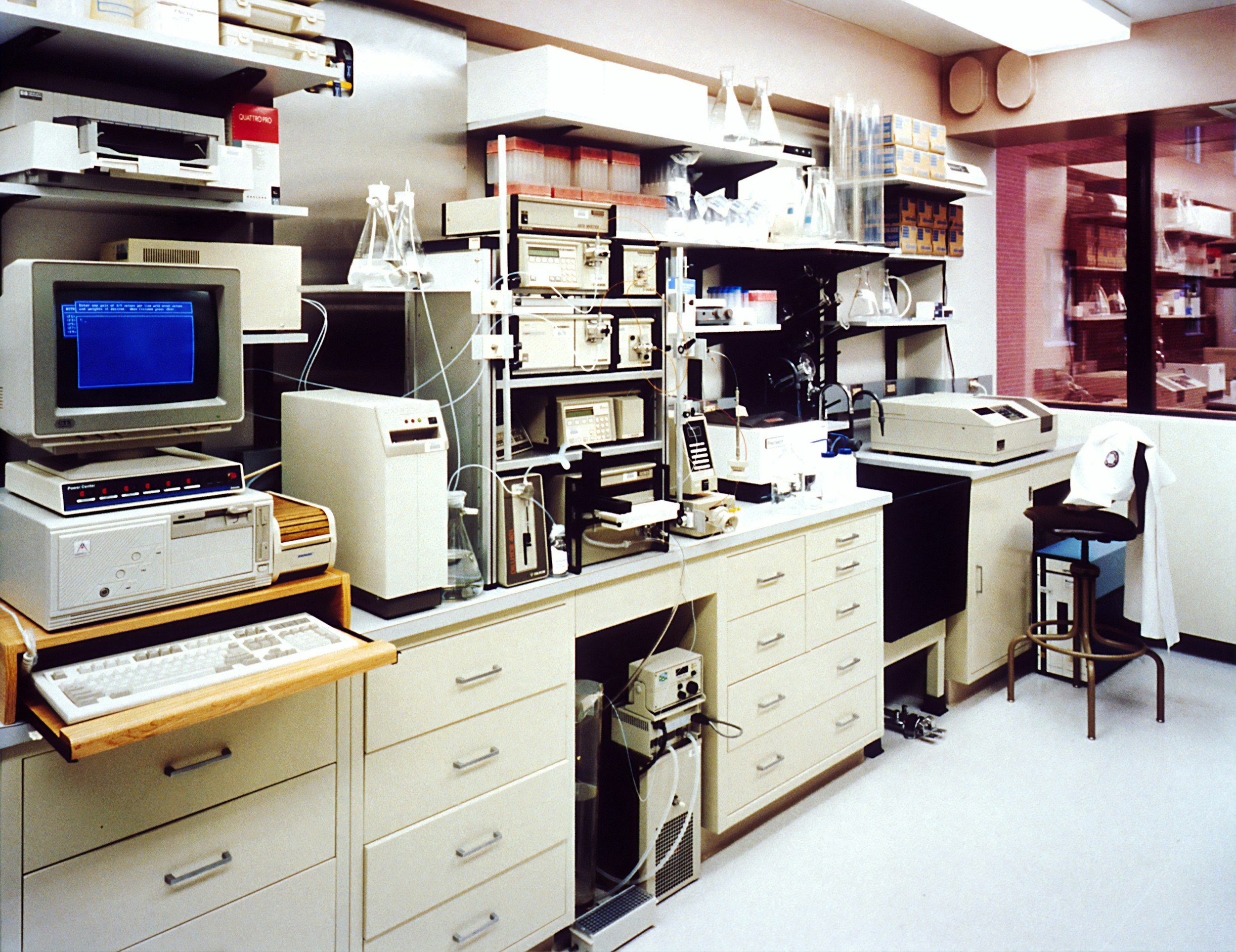 Laboratory with scientific equipment including monitors, centrifuges, microscopes, and glassware on shelves and countertops.