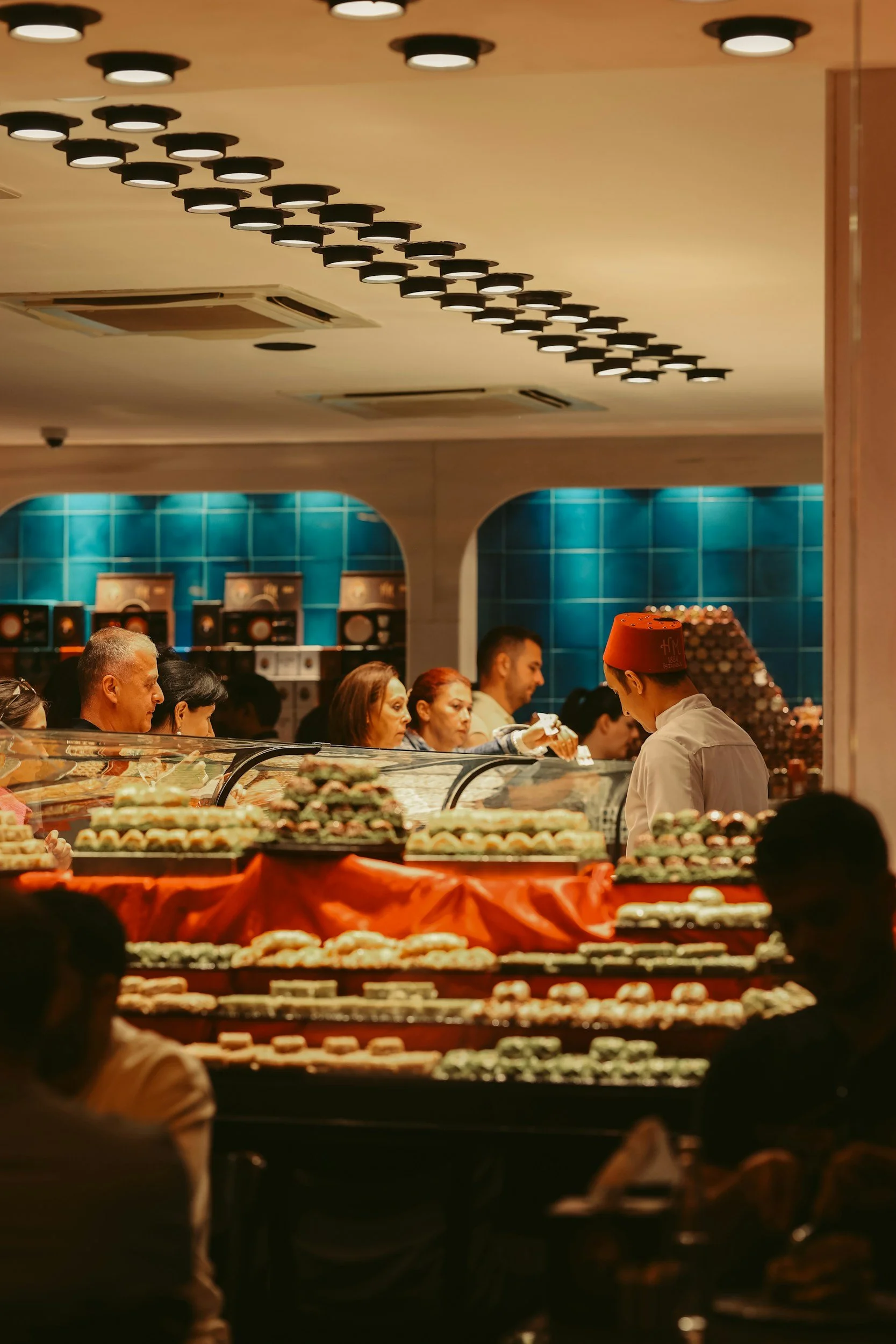 People waiting in line at a bakery counter with various pastries on display, inside a well-lit bakery or dessert shop.