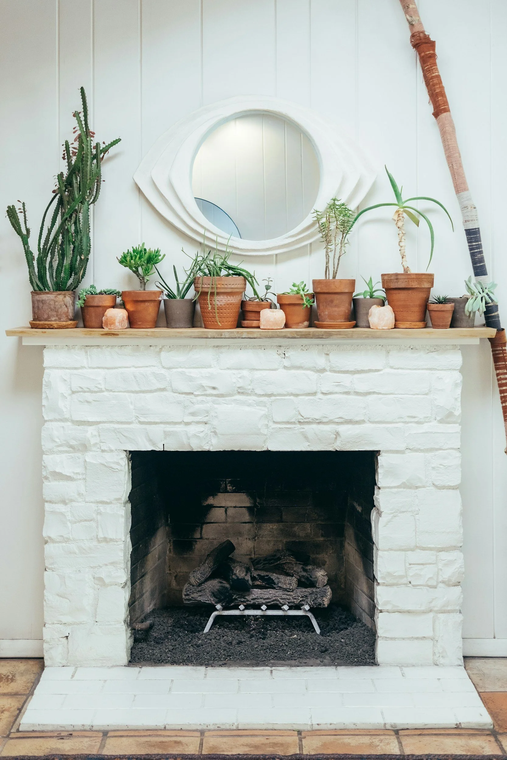 White brick fireplace with a wooden mantle decorated with potted plants and a circular mirror above.