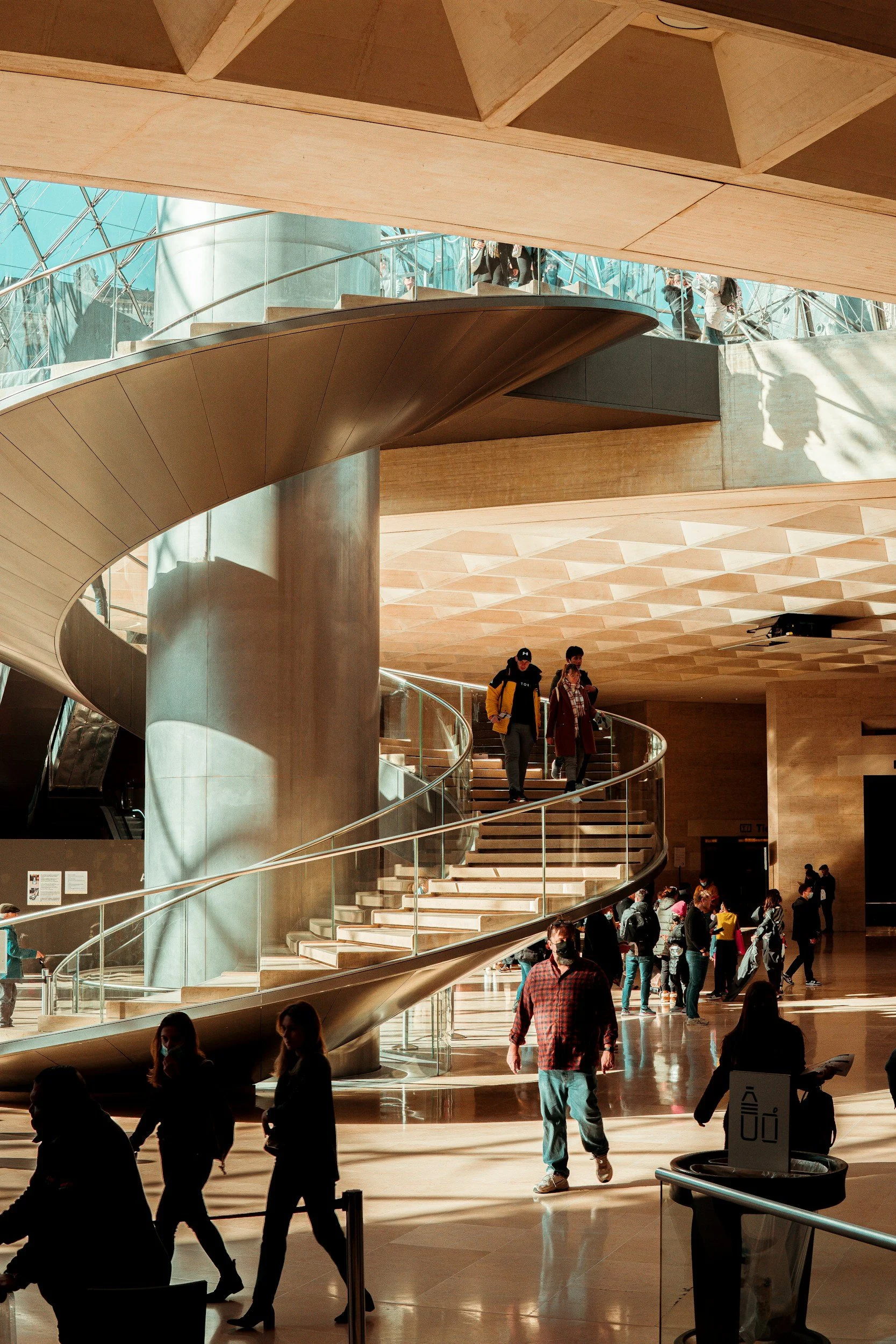 People walking and descending a spiral staircase in a modern, spacious building with natural light and wooden interior design.
