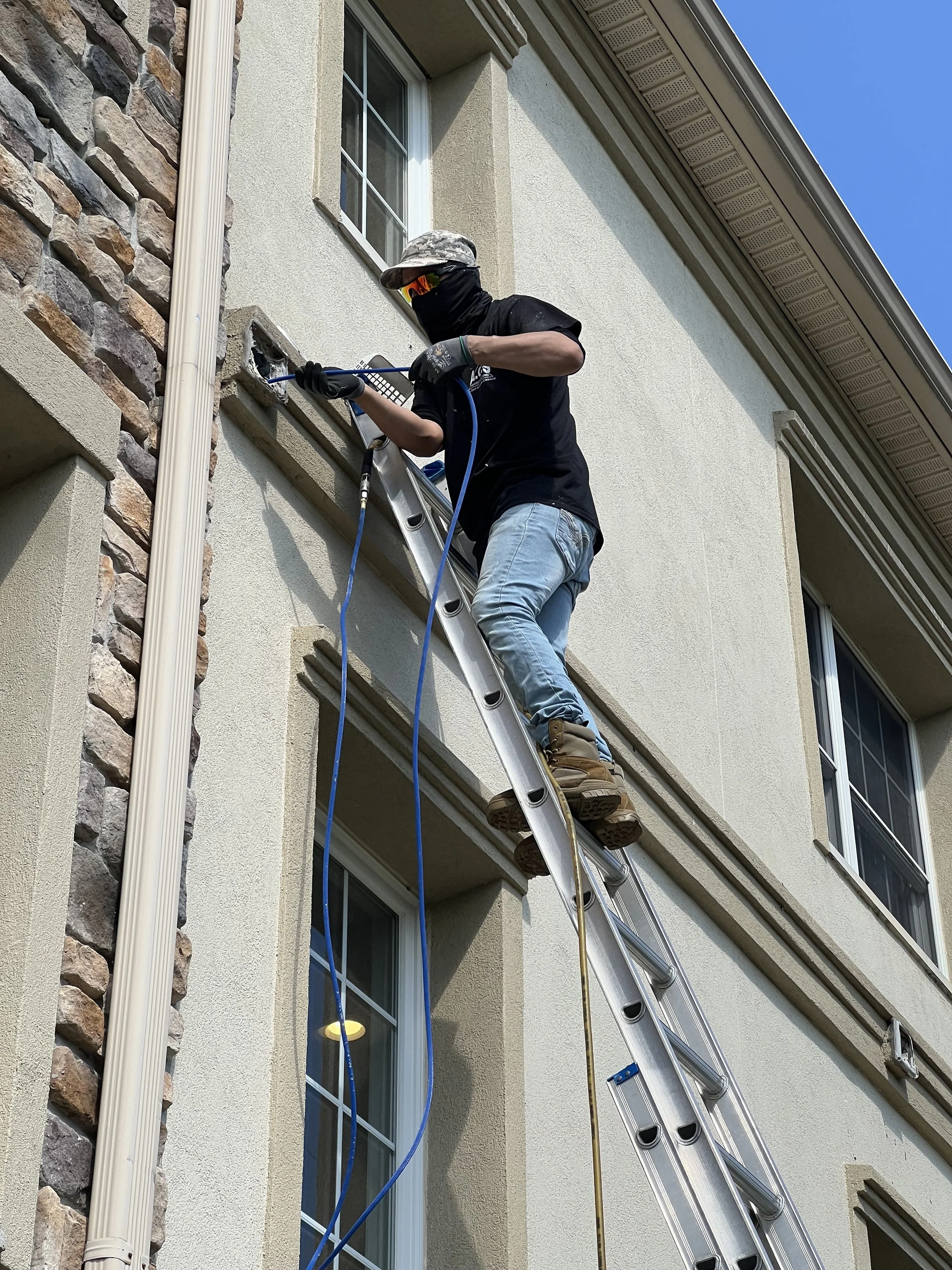A person standing on a ladder working on the exterior of a house, wearing a mask, gloves, and a hat, with tools in hand.