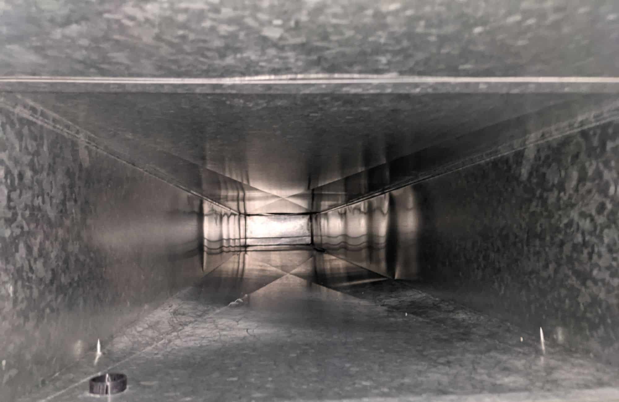 View inside a metal duct or ventilation shaft showing reflections and an opening at the end.