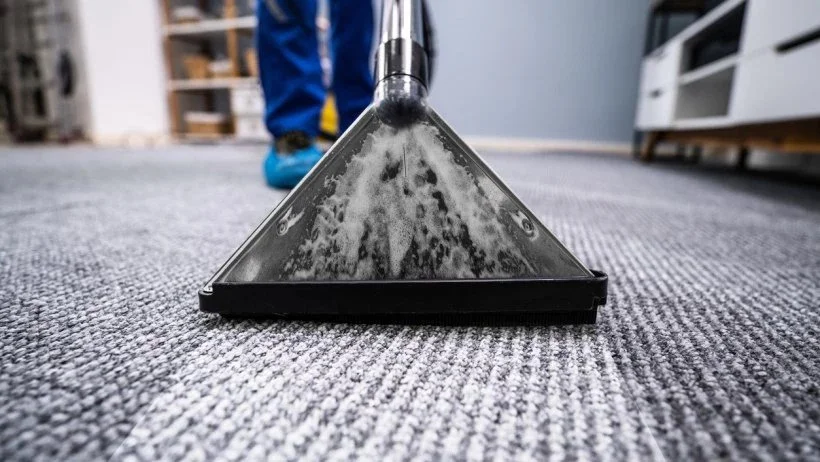 Close-up of a carpet cleaning tool spraying water and cleaning solution on a gray carpet in a room with shelves and a person in the background.