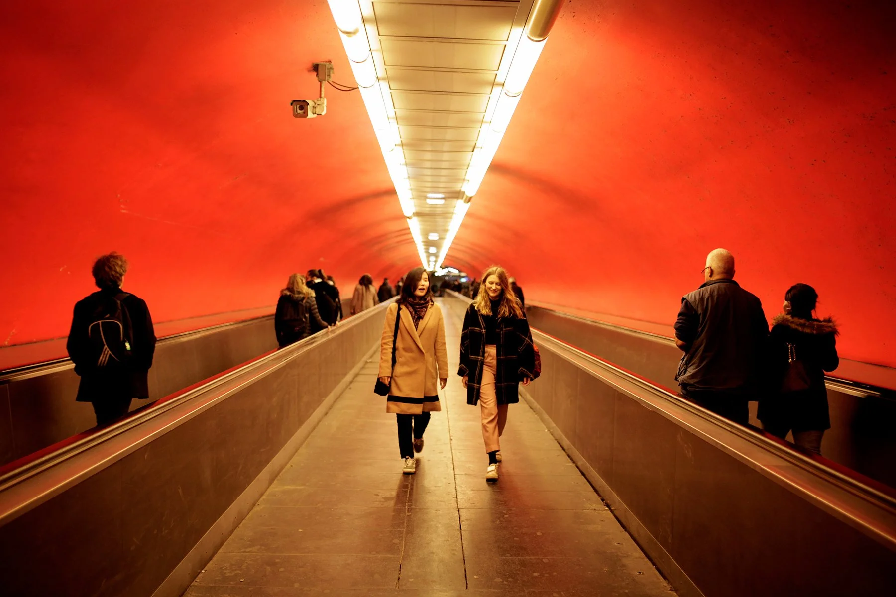 People walking through an underground tunnel with bright lighting and reddish-orange walls.