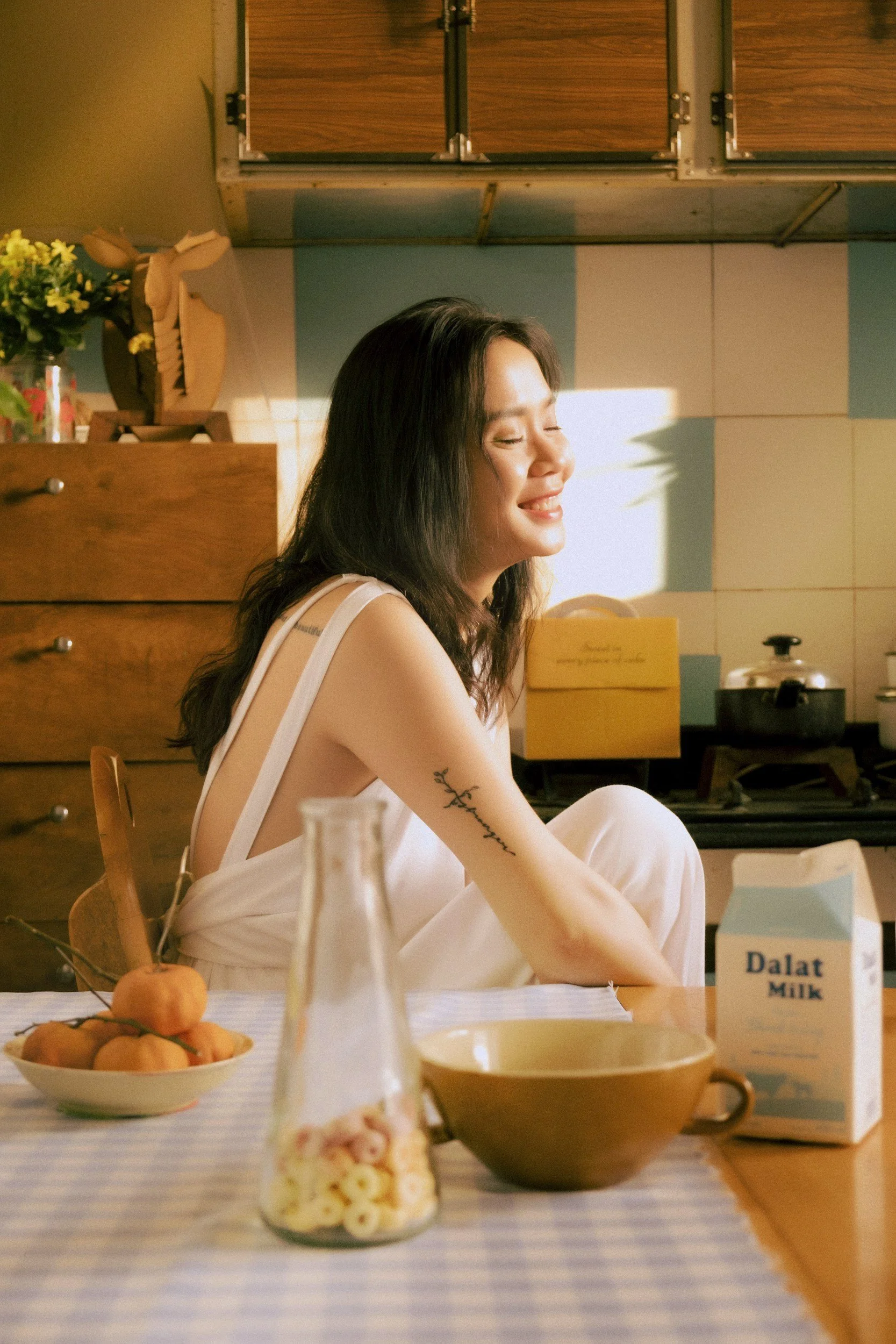 A woman sitting at a dining table with her eyes closed and a smile, wearing a white dress, in a kitchen with wooden cabinets and a tiled wall. The table has a bowl of peaches, a glass jar of cereal, a brown mug, a carton of milk, and a plate with a spoon. Sunlight is shining on her face.