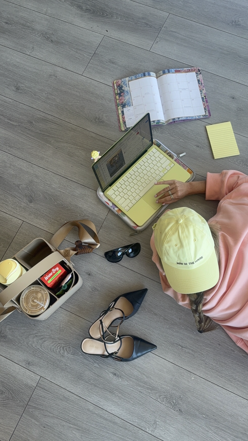 A person with a yellow cap and pink sweatshirt sitting on a wooden floor surrounded by a laptop, a planner, sunglasses, a pair of black shoes, a lunch box with a water bottle, and a yellow notepad.