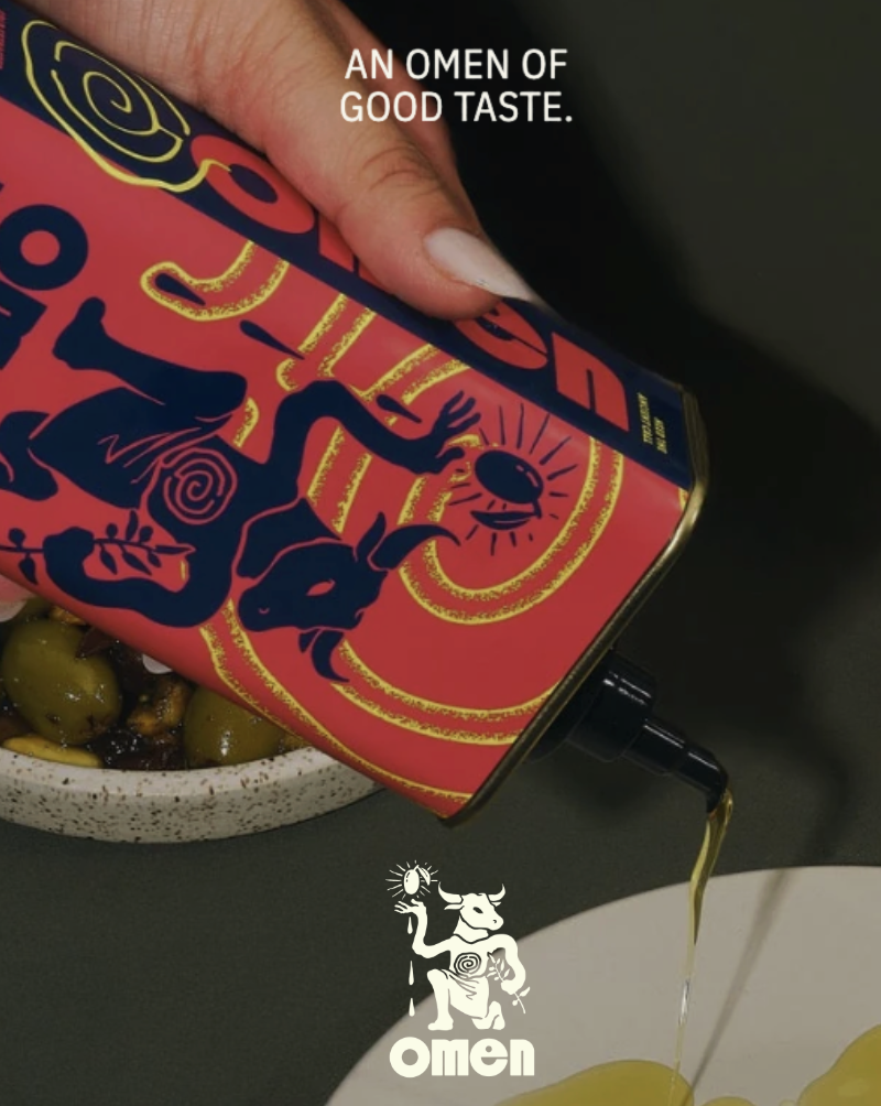 A hand pouring olive oil from a decorative red and blue canister with a mythical creature illustration onto a plate of green and black olives. The canister is branded "Omen" and has the slogan "An omen of good taste."