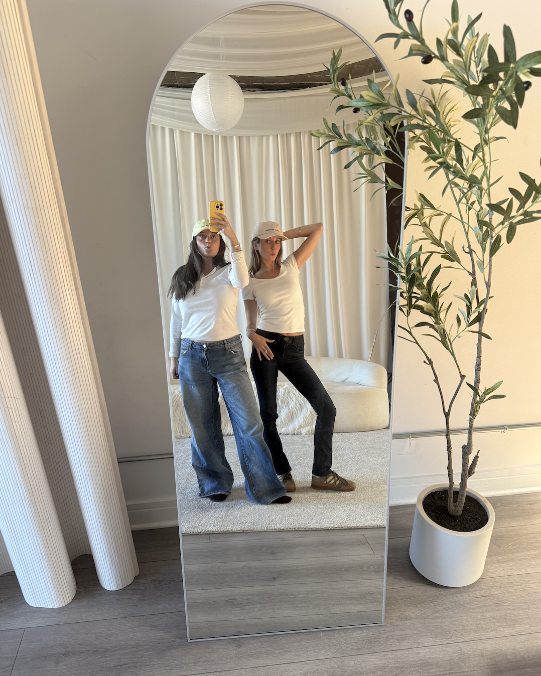 Two women taking a mirror selfie in a modern room with a tall potted olive tree, white curtains, a round white ceiling light, and light wooden floors. One woman is wearing a white long-sleeve top, wide-leg jeans, and a yellow cap. The other woman is in a white t-shirt, black jeans, and a white cap.