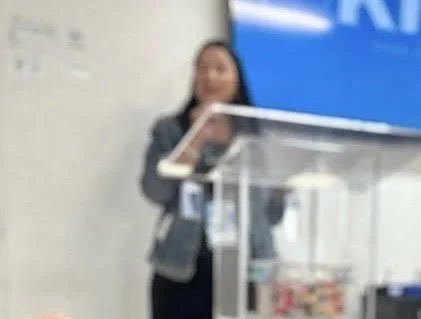 A woman standing behind a glass display case in a retail or pharmacy setting.