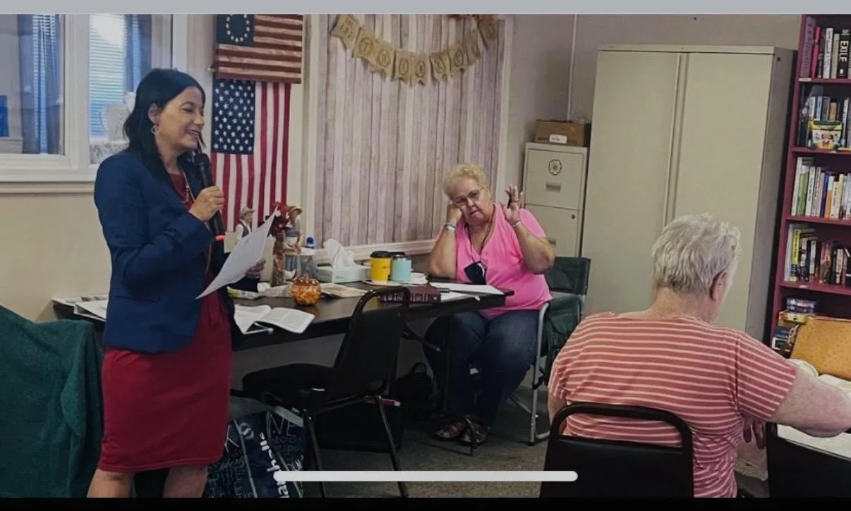 A woman in a navy blue blazer and red dress standing and speaking into a microphone in a room with American and European Union flags and a 'Graduation' banner on the wall, with three senior women sitting at tables listening.