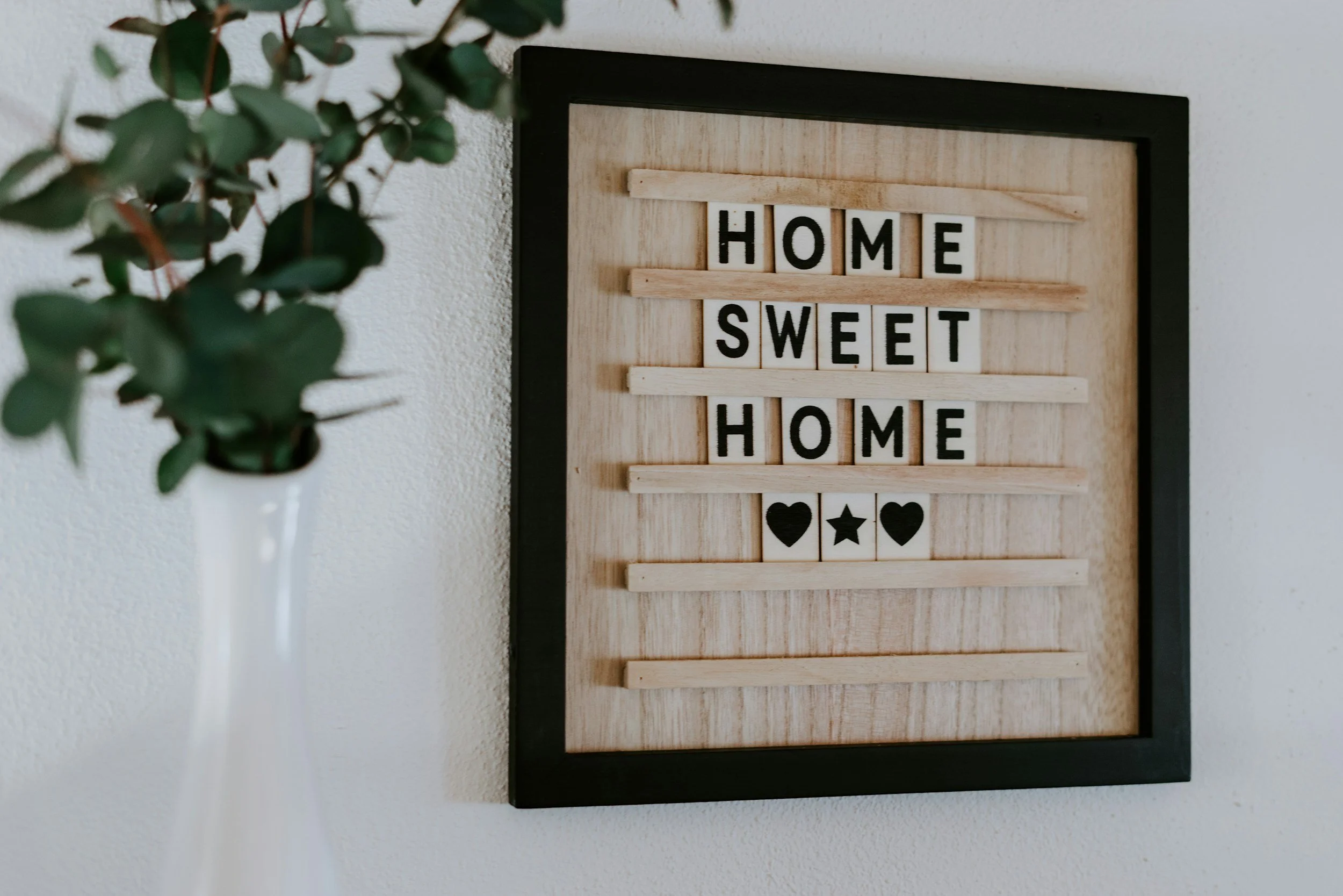 A framed decorative sign on a white wall with the words 'Home Sweet Home' in black letters and three small icons (heart, star, heart) below, next to a white vase with green leafy plant.
