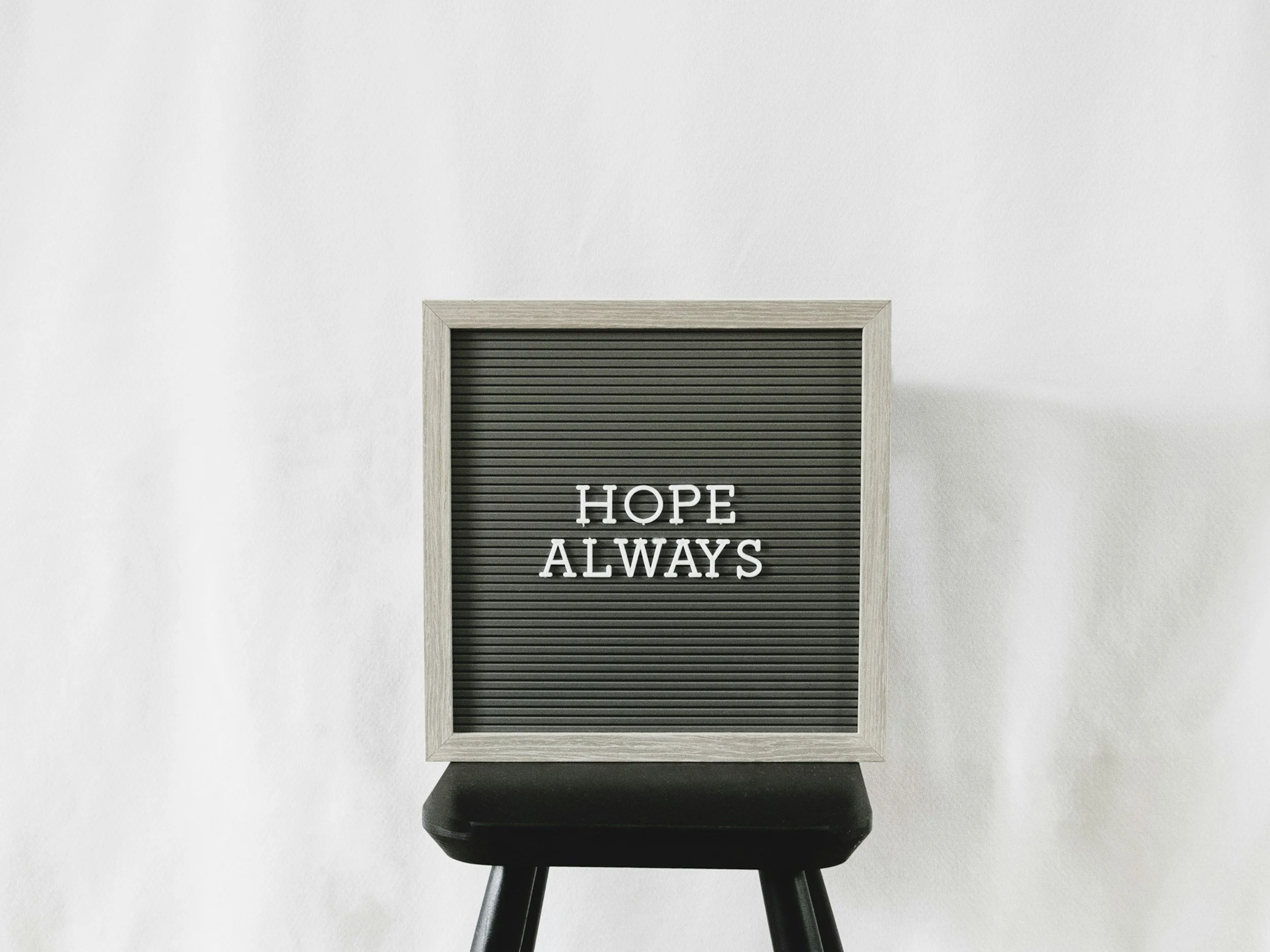 A black felt letter board with the message "HOPE ALWAYS" placed on a black stool against a white wall.