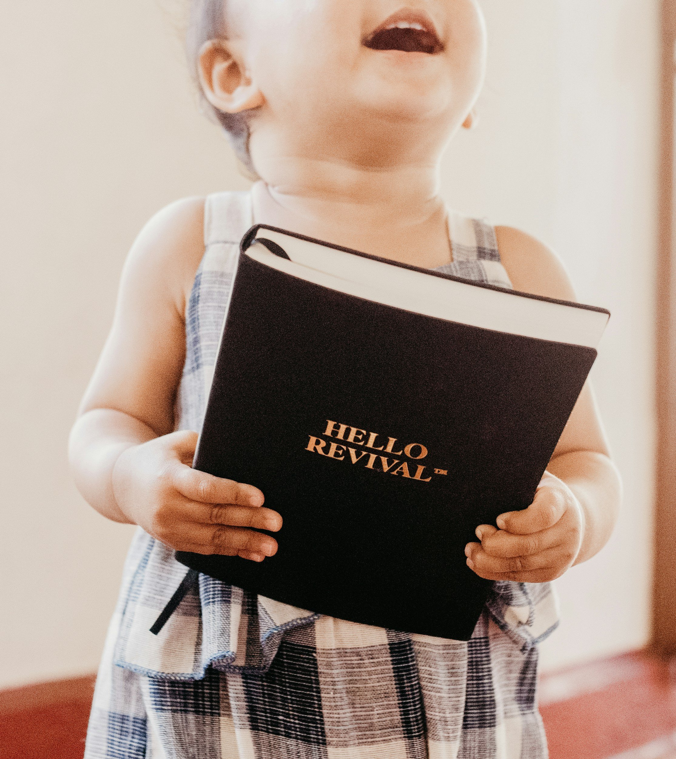 A young child holding a black notebook with the text 'HELLO REVIVAL' on the cover, wearing a sleeveless checkered outfit, indoors.