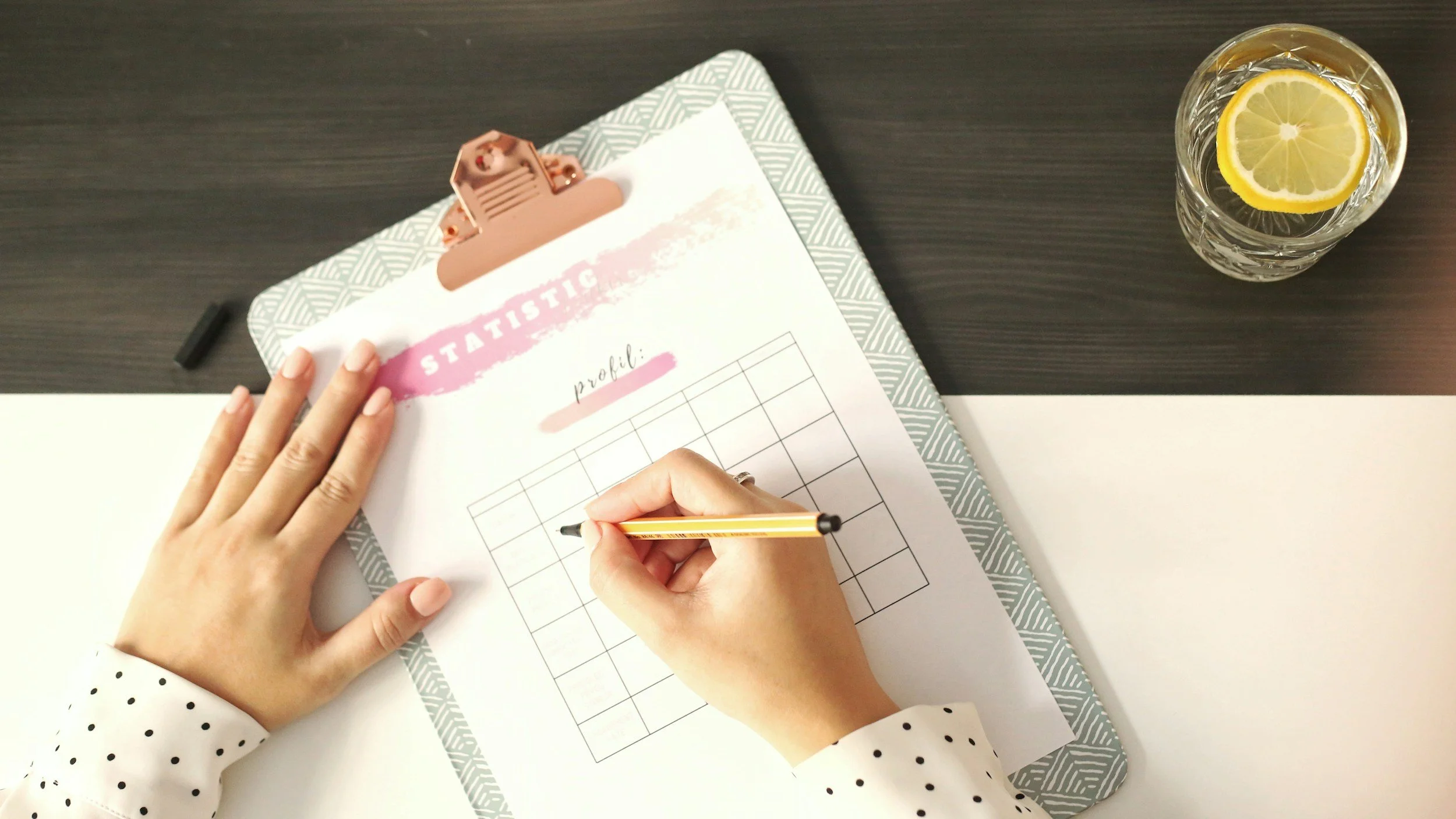Person filling out a spreadsheet titled 'Statistics' with a yellow pen on a clipboard, with a glass of water and lemon slices on the desk.