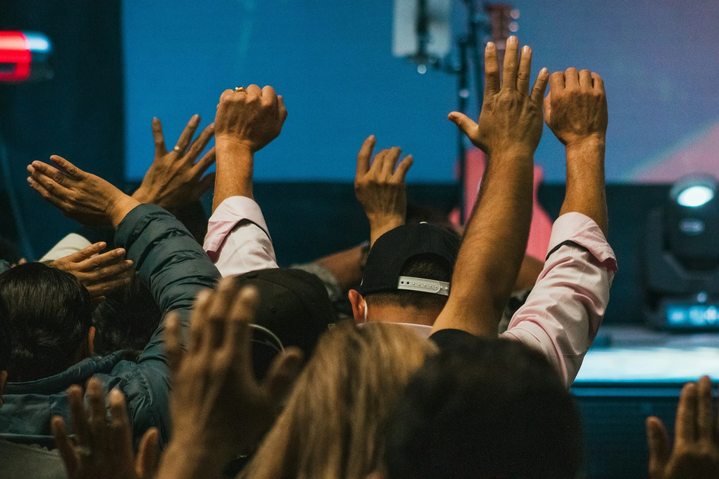 A large crowd of people raising their hands, some with rings, in an indoor setting with blue and pink lighting.