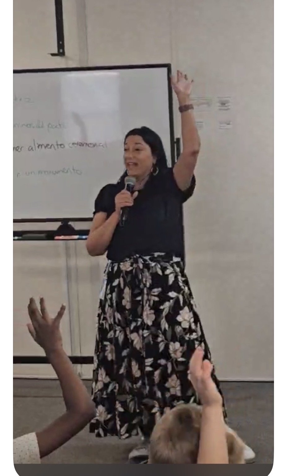 A woman standing in front of a whiteboard, holding a microphone, raising her right hand, and speaking to an audience with raising hands and a dog in the foreground.