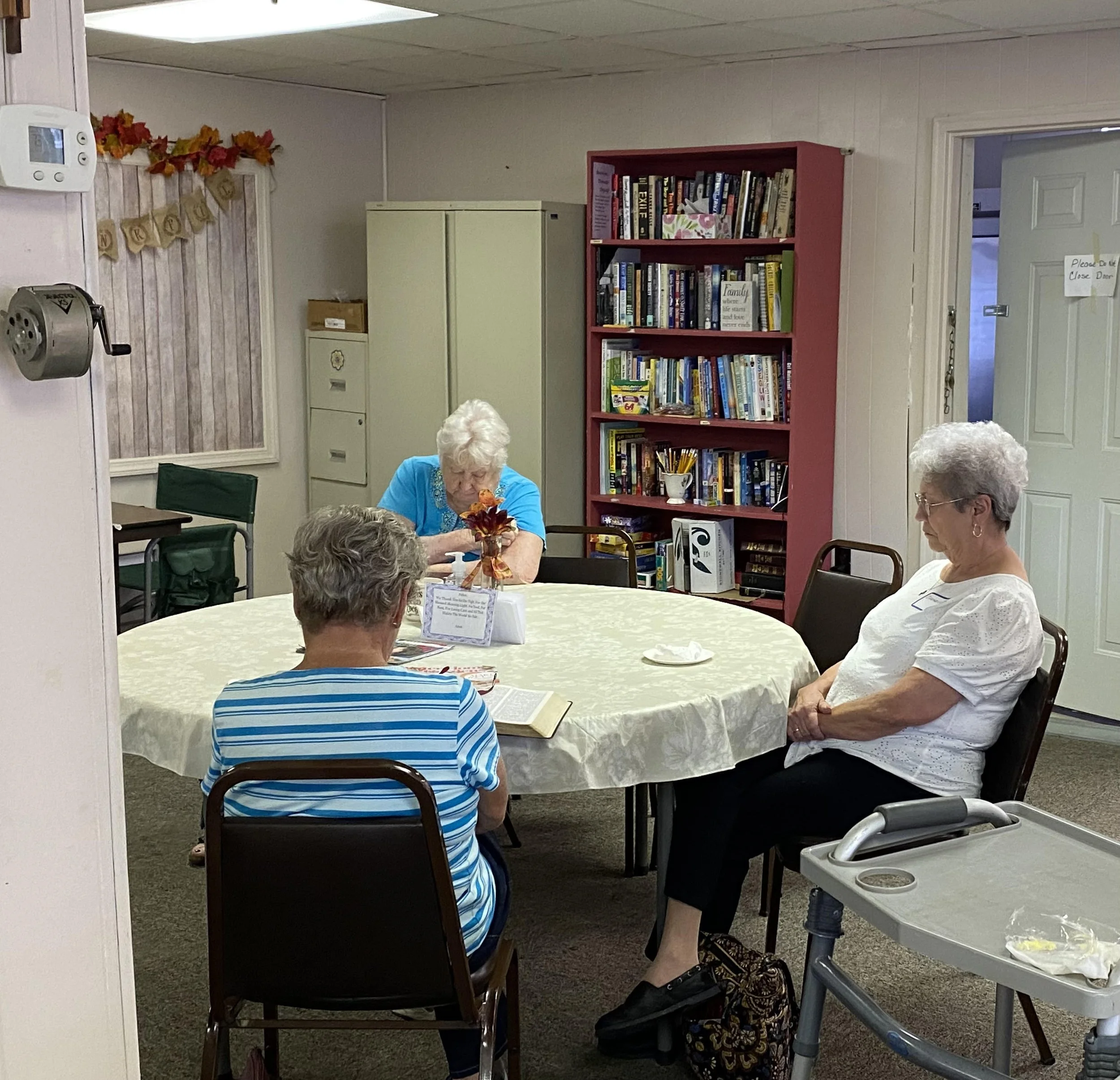 A group of elderly women sitting around a table in a room with a bookshelf, a cabinet, and a wall decorated with autumn leaves and a banner. One woman in a blue shirt is looking down at a book, others are engaged in conversation.