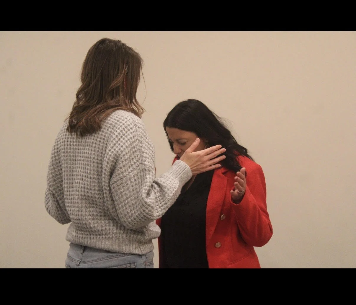 Two women, one with brown hair and the other with black hair, are engaged in a close interaction against a plain background. The woman with brown hair is wearing a gray sweater and touching the face of the woman with black hair, who is wearing a red jacket.