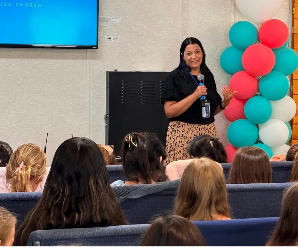A woman holding a microphone and speaking to an audience of children indoors near colorful balloons and a large screen.