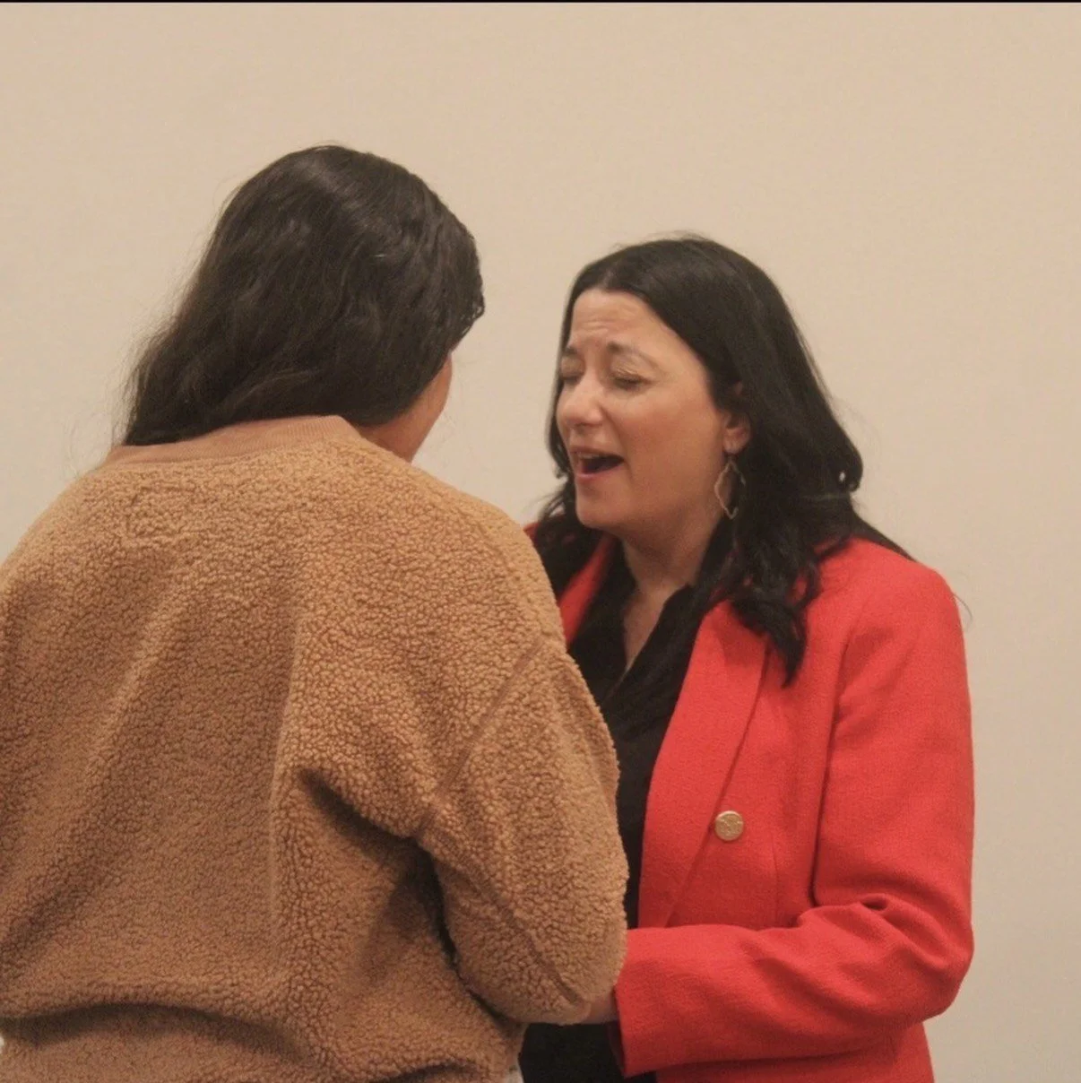Two women are engaged in a conversation; one has long dark hair and is wearing a brown fuzzy jacket, and the other has black hair, earrings, and is wearing a red blazer.
