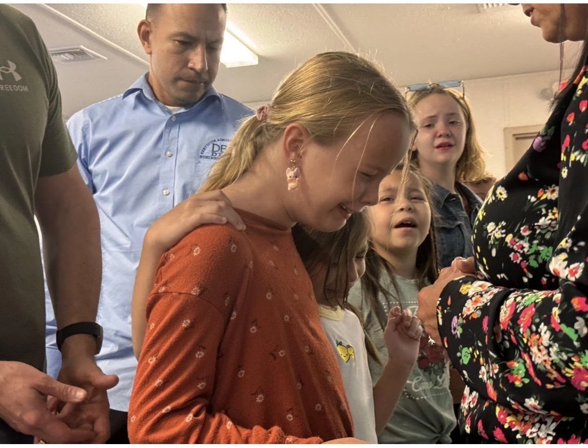 A group of children and adults gathered together, with a girl in an orange sweater smiling and looking down, while others look on with varied expressions.
