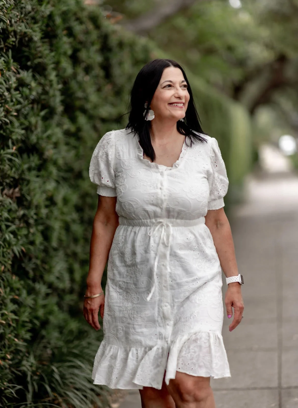 A smiling woman with black hair in a white dress walking outdoors on a pathway lined with greenery.