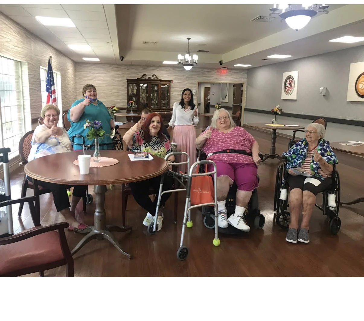 Group of seven women, some in wheelchairs, in a decorated dining or activity room, smiling and giving thumbs-up gestures.
