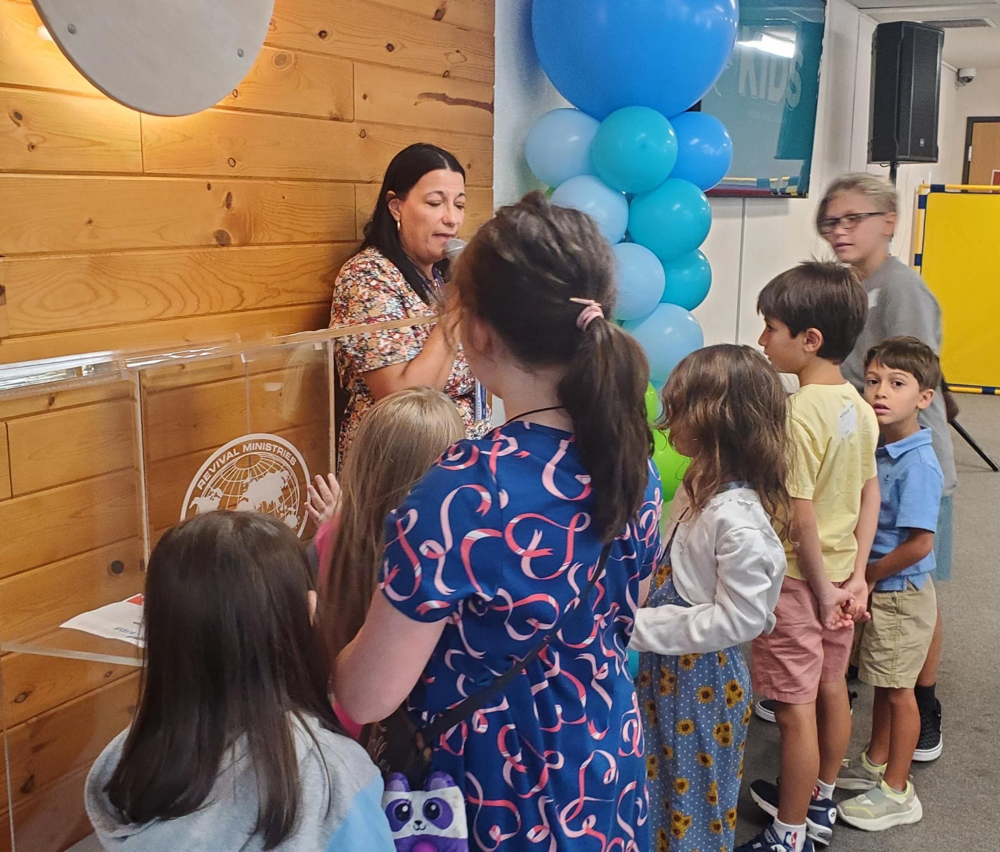 A group of children gathered around a woman with dark hair who is speaking into a microphone, with blue balloons and wooden wall in the background, likely at a celebration or event.