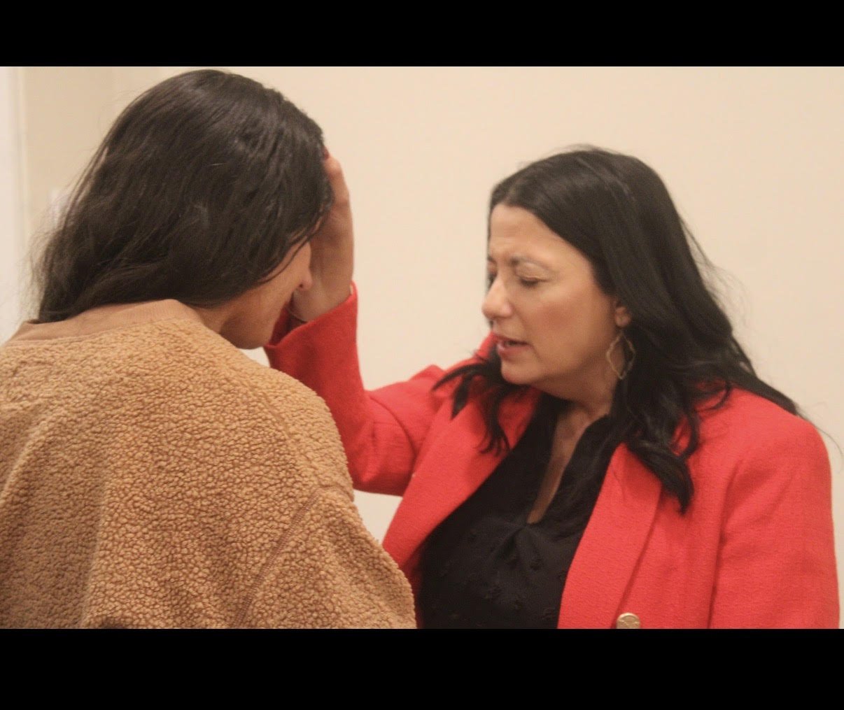 Two women, one in a beige coat and the other in a red blazer, sharing an emotional moment with the woman in the red blazer comforting the other woman who is distressed.