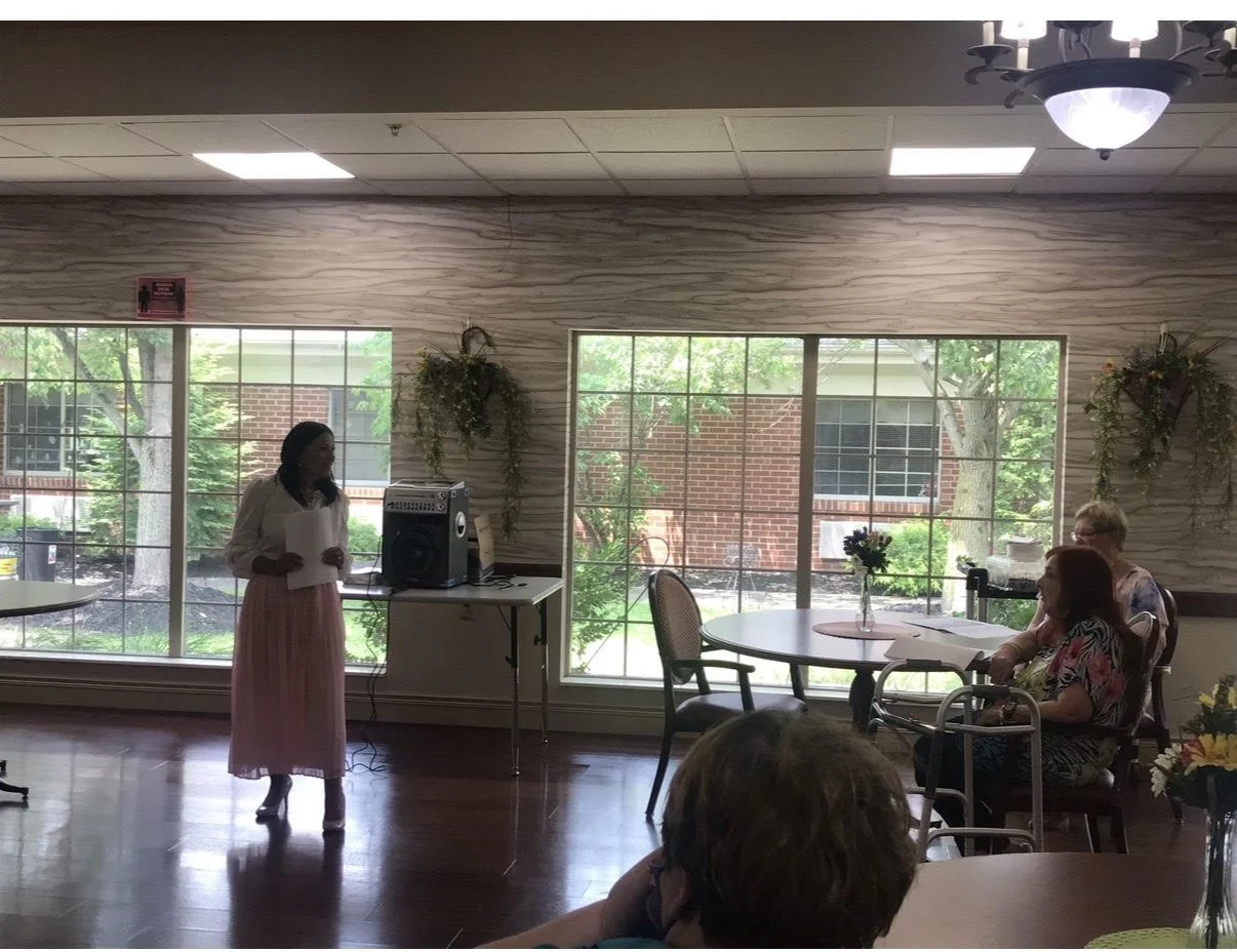A woman standing near a window and speaking into a microphone during a gathering with seated attendees.
