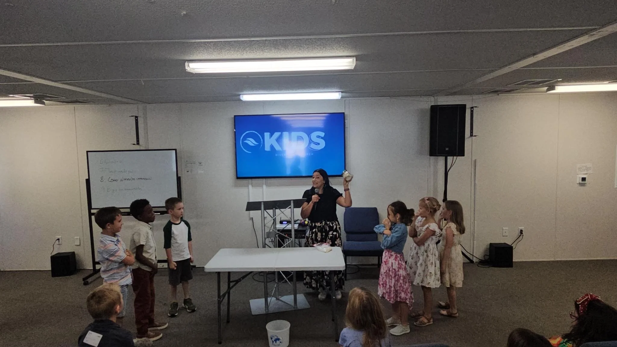 A woman leading a children's activity in a classroom or community room, with six children standing in front of her. The woman is smiling and holding a globe. There is a large screen behind her with the word 'KIDS' on it.