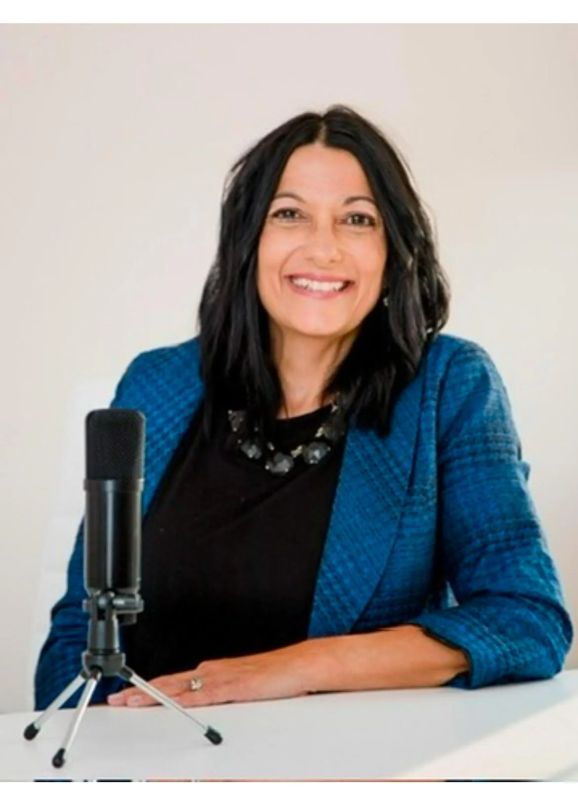 A woman with black hair and a bright smile, wearing a blue blazer and black top, sitting in front of a white background with a small microphone on a tripod in front of her.