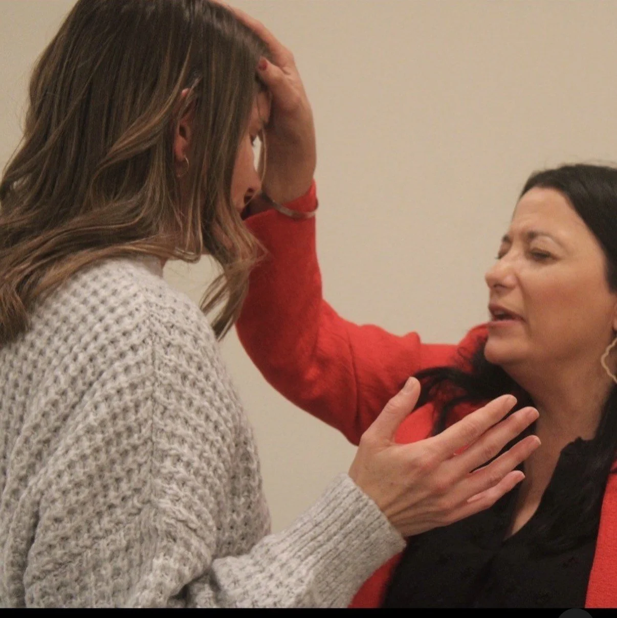 Two women are in an emotional interaction. The woman on the right, dressed in red, appears to be comforting or speaking passionately to the woman on the left, who is in a white sweater. The woman on the left has her hand on her forehead and appears distressed.
