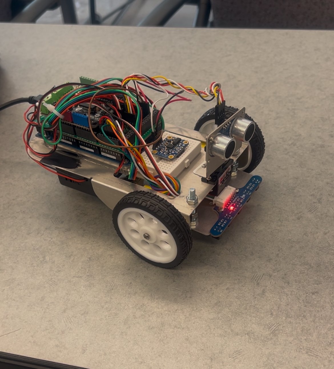 A small robotic vehicle with wires, sensors, and electronic components on a beige table.