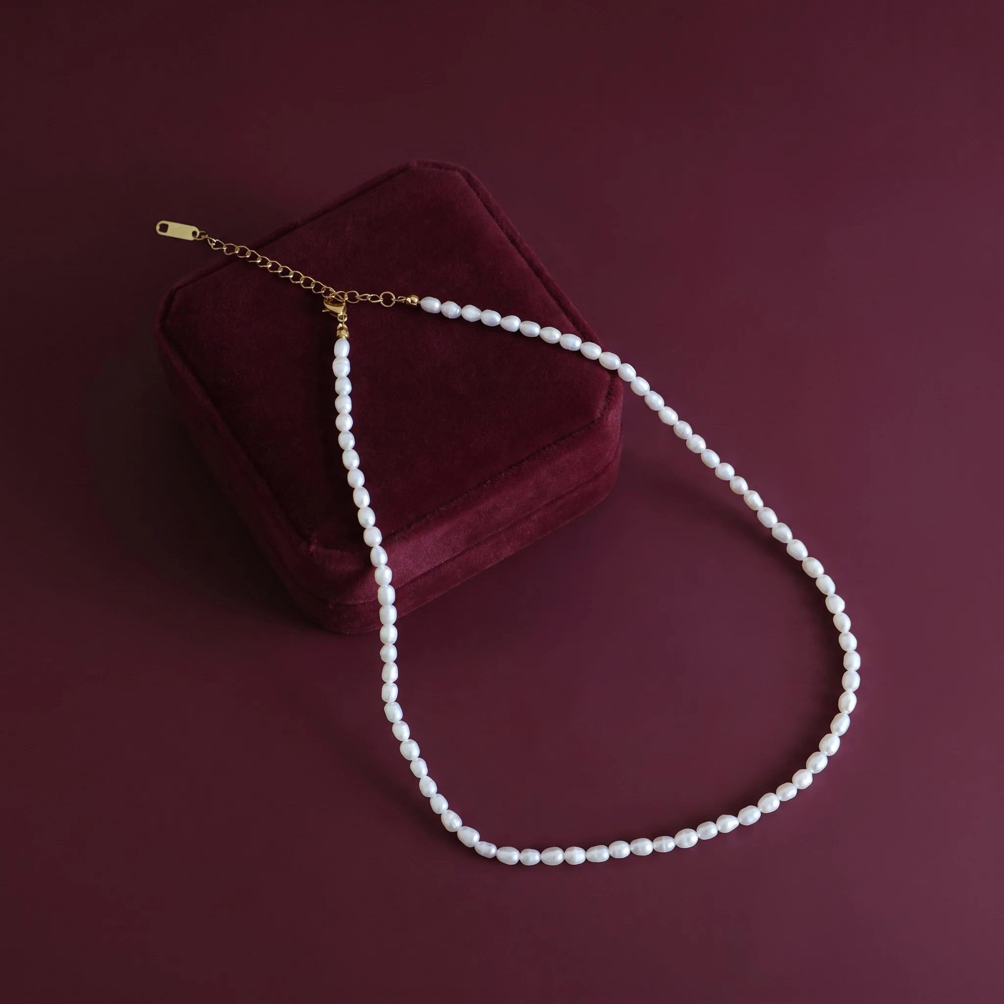 Delicate natural freshwater rice pearl choker necklace arranged beside a burgundy velvet jewellery box with gold-plated clasp and extension chain.