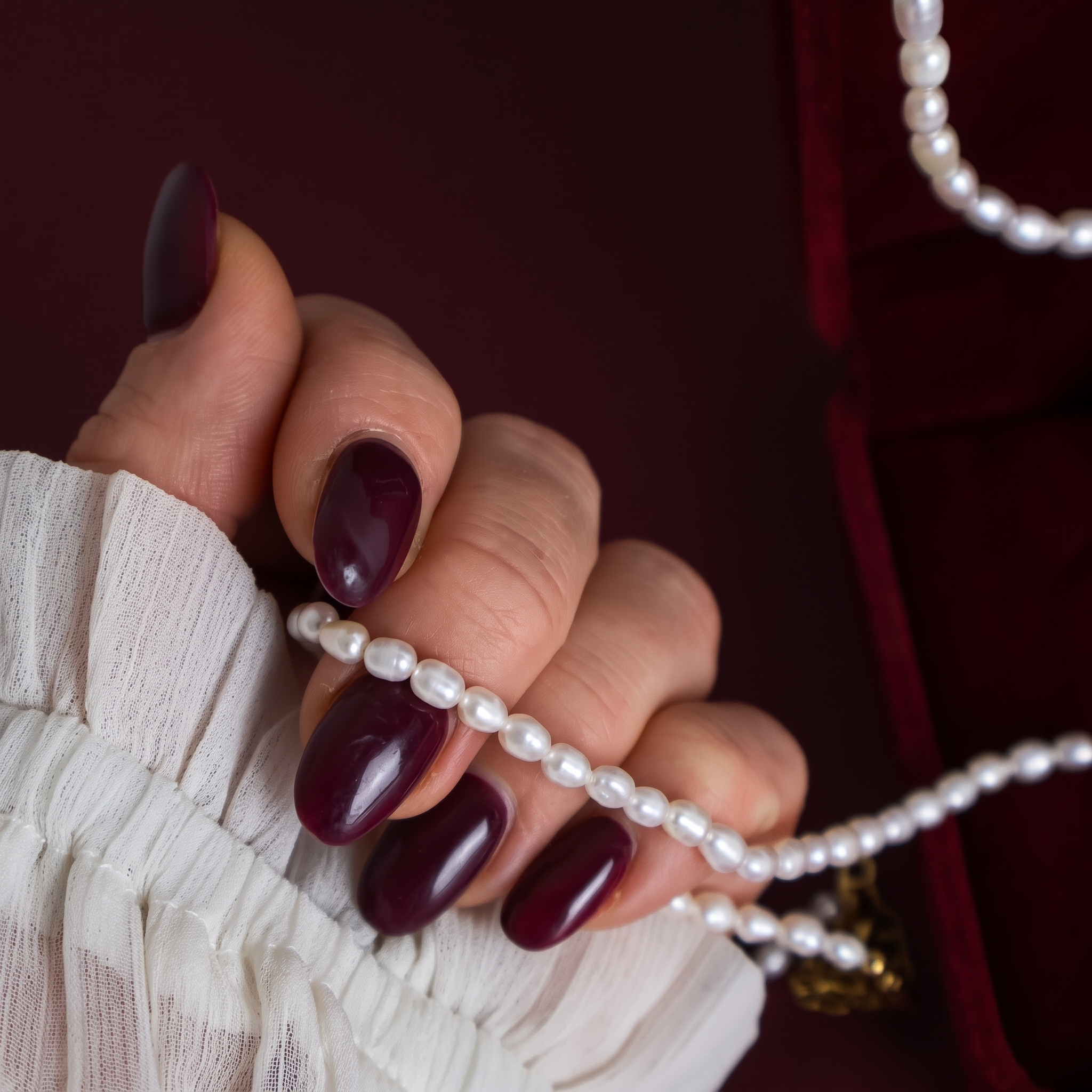 Close-up of a hand with burgundy manicure holding a natural freshwater rice pearl necklace, highlighting the pearls' shape and lustre.