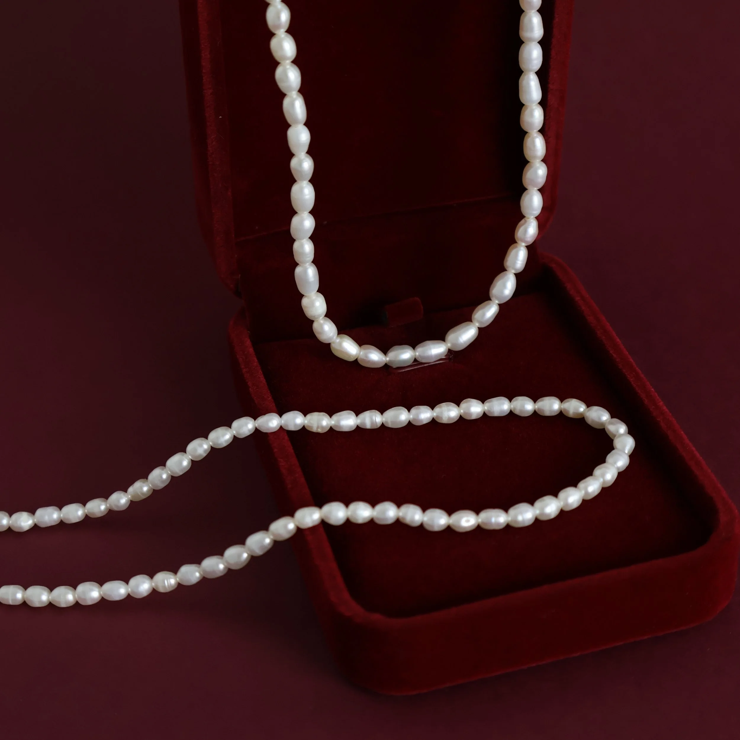 Natural white freshwater rice pearl necklace draped over a deep burgundy velvet jewellery box, showing the delicate oval pearls.