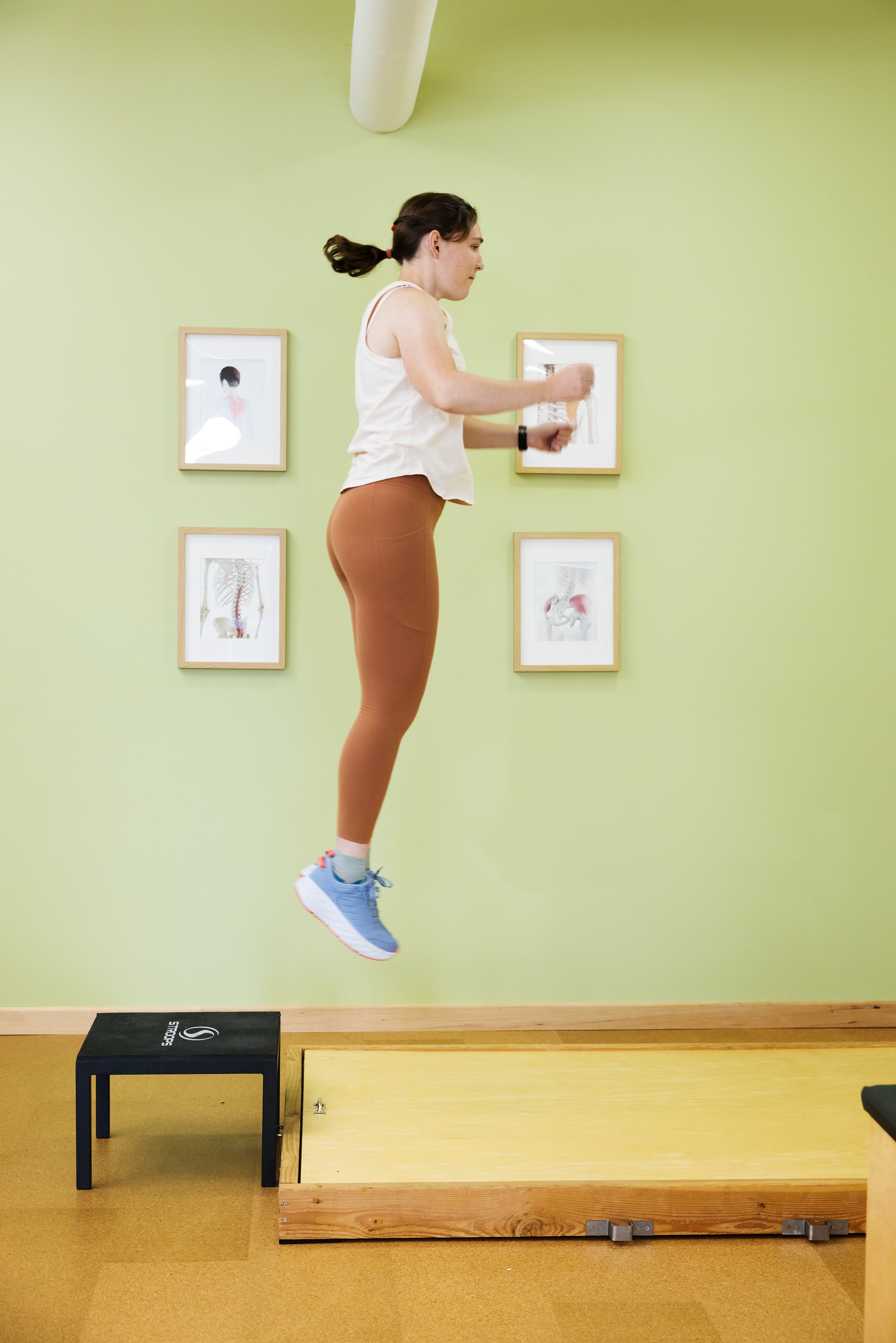 Vertical photo of a woman in athletic attire mid air, jumping off a short black stool onto a small wood platform with a in a calm space with light green walls at ActiveLab gym in Vancouver, WA