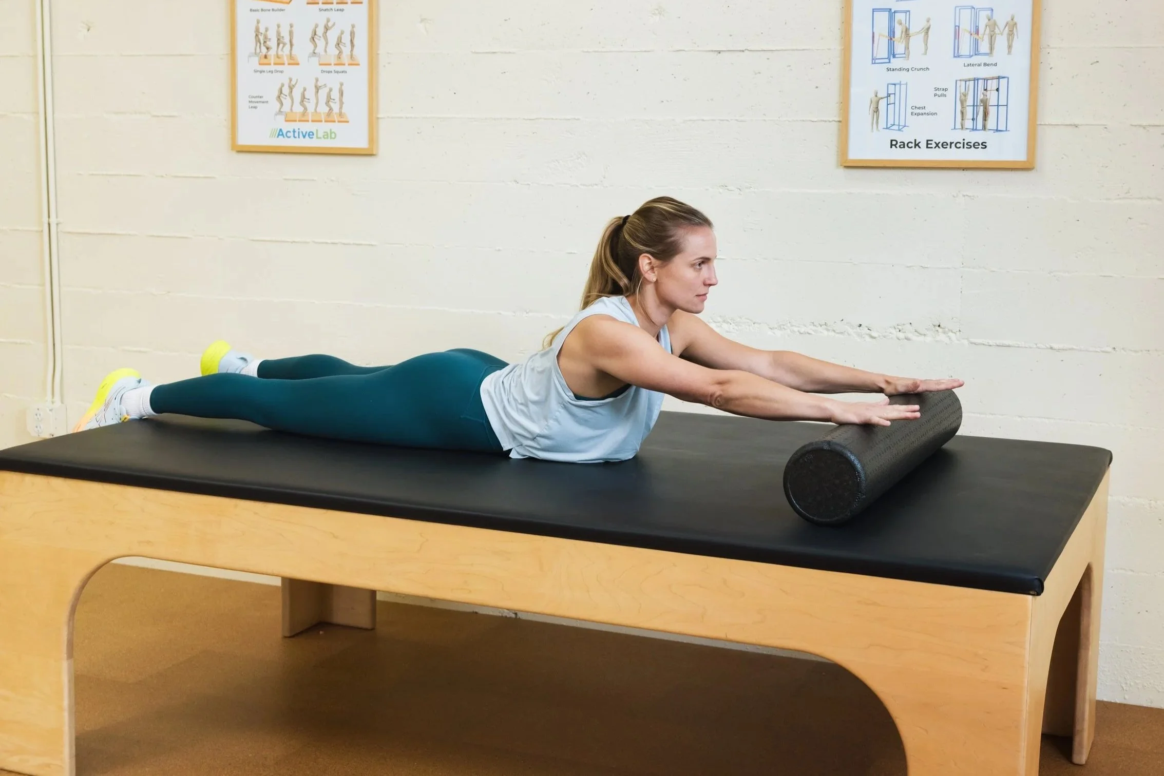 Photo of a woman in athletic attire, lying belly-down on a padded exercise table at ActiveLab gym in Vancouver, WA, using a foam roller under her hands and wrists to roll out and stretch her spine
