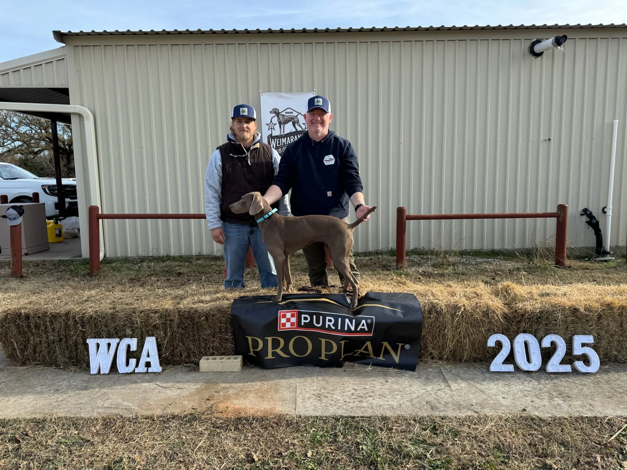Two men standing outdoors in front of a beige metal building, smiling, with a brindle dog on a leash. There are decorations including white letters spelling 'WCA 2025', a black banner with the 'Purina Pro Plan' logo, and white letters spelling 'TWCA'. One man wears a black hoodie, and the other wears a vest and hoodie. Both wear caps. The scene appears to be at a dog event or competition.