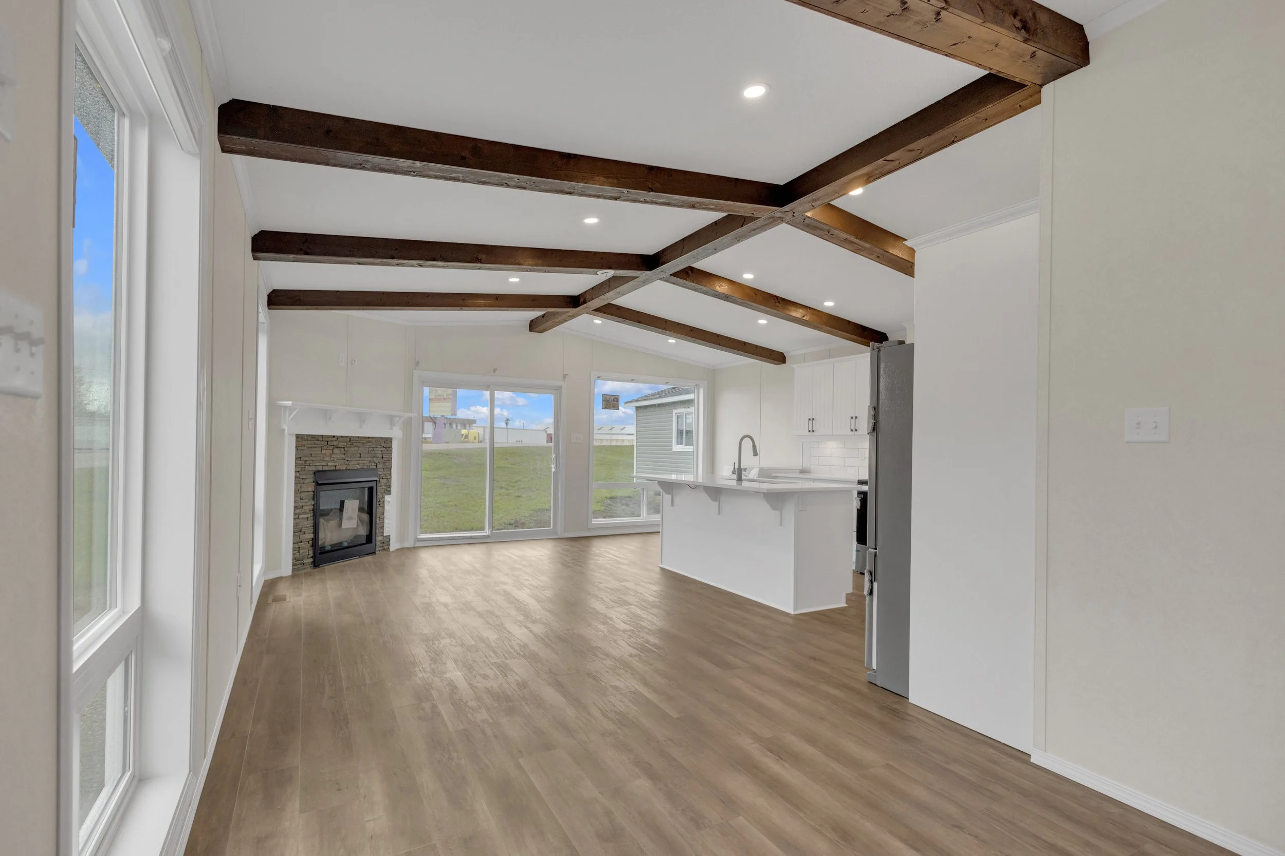 An open living room and kitchen area with large windows, wooden ceiling beams, a fireplace, and laminate flooring.
