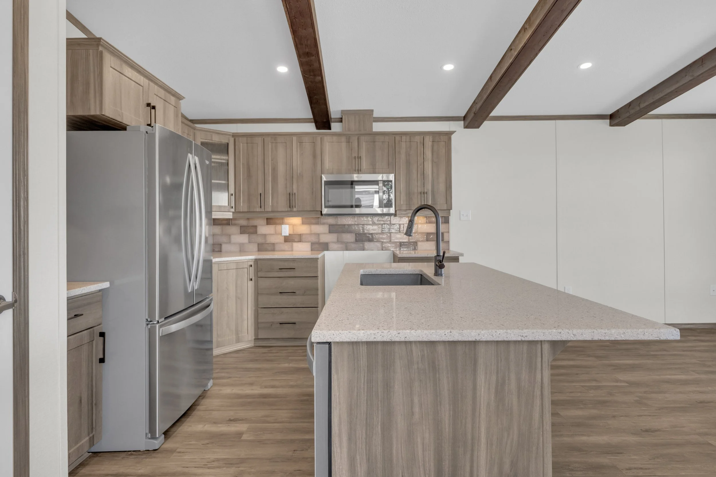 Modern kitchen with wooden cabinets, stainless steel refrigerator, microwave, brick-style backsplash, and a large island with a sink and faucet, wooden beams on the ceiling, and wood flooring.