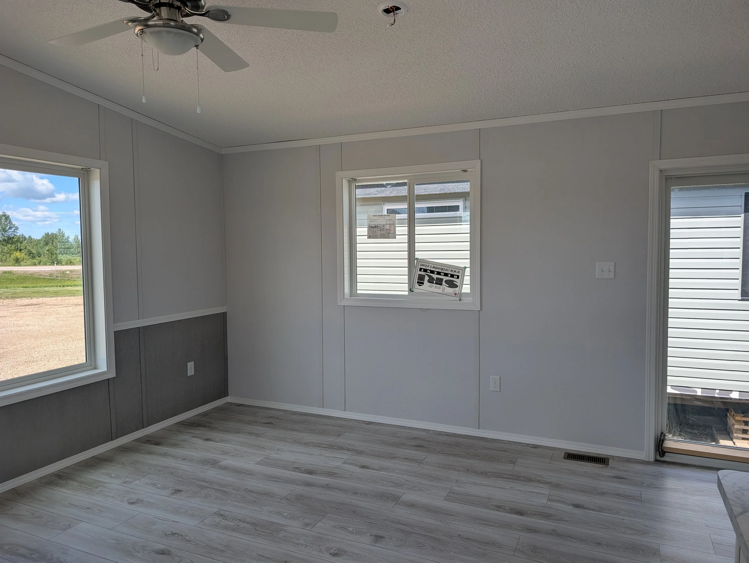 An empty room with light-colored wood flooring, white paneled walls, three windows letting in natural light, and a ceiling fan with a light fixture.