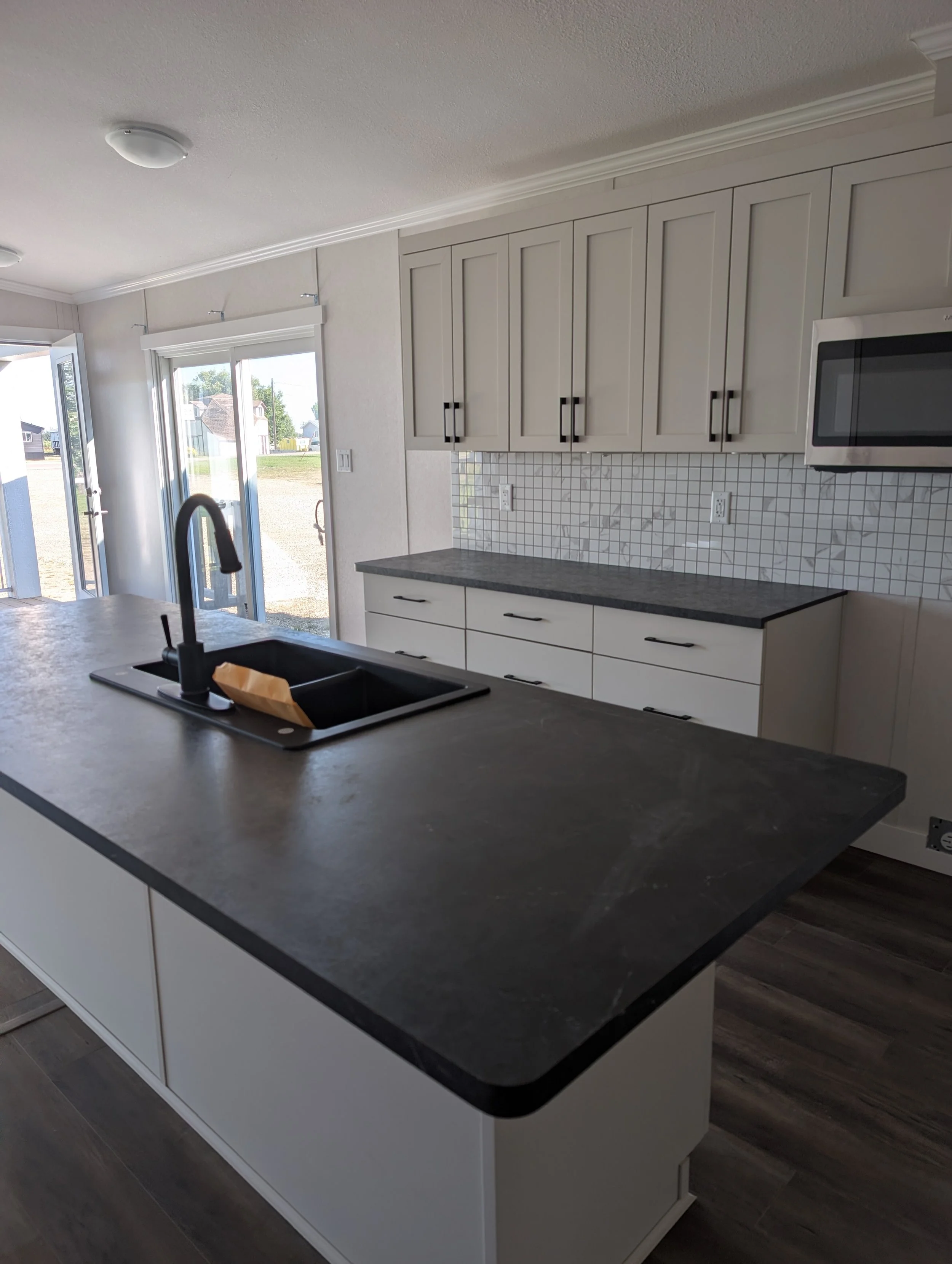 Clean modern kitchen with gray countertops, white cabinets, tile backsplash, black handles, stainless steel microwave, sink, cabinets, sliding glass door with outside view, and wood flooring.