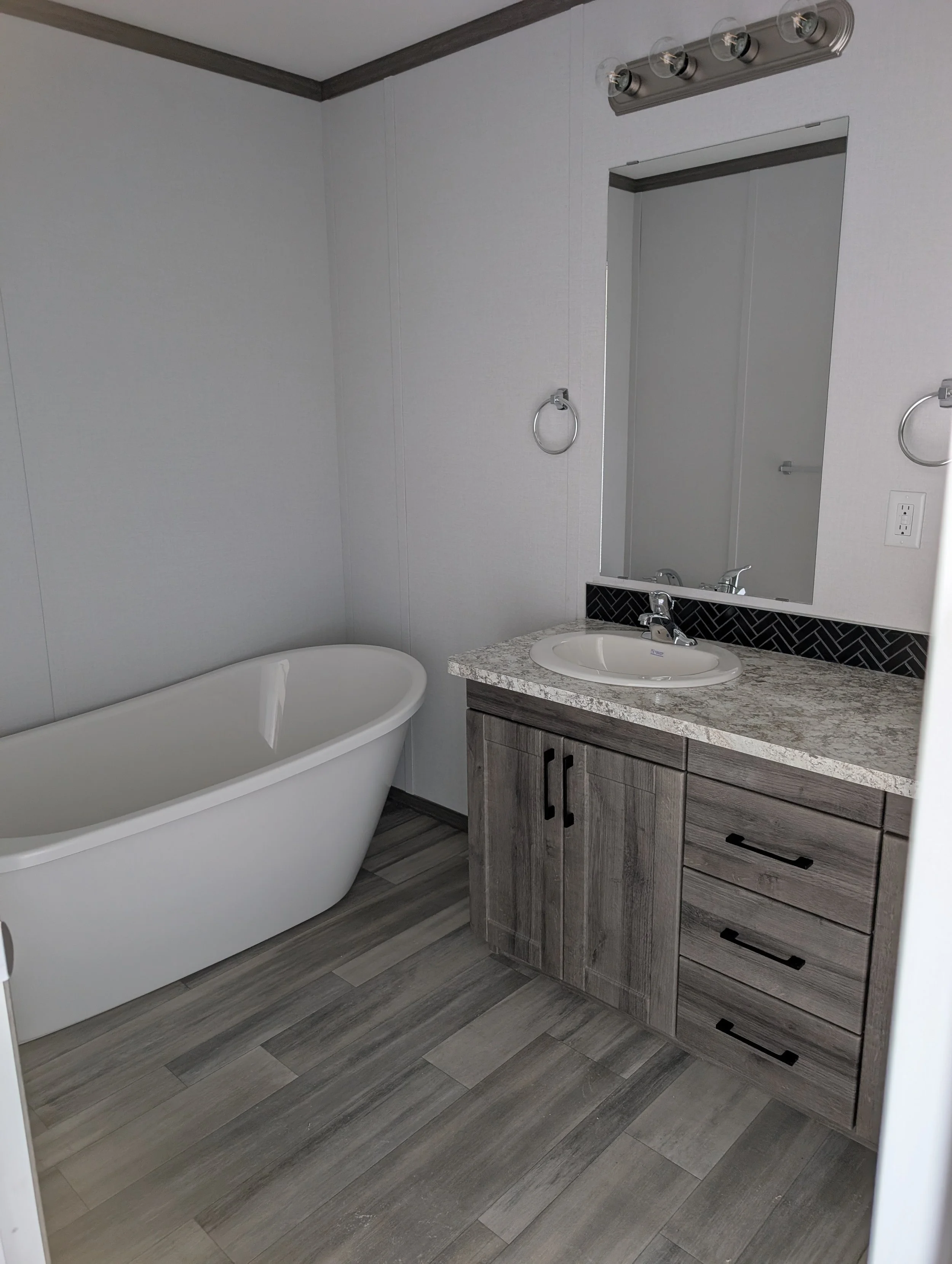 A bathroom with a white bathtub, a vanity with a granite countertop, a mirror, and a row of four exposed light bulbs above the mirror. The walls are white, and the floor and cabinetry are in shades of gray.